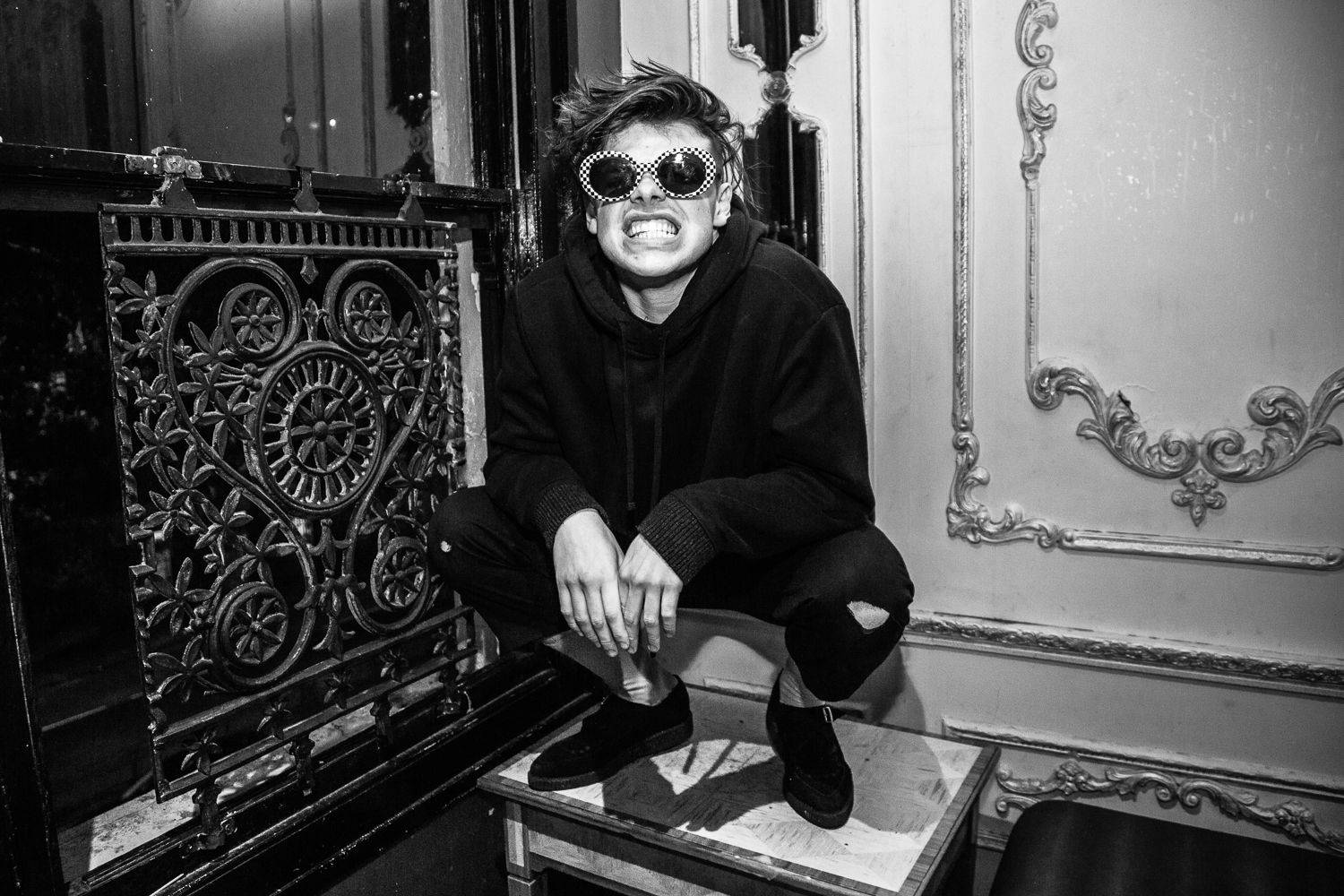 Yungblud 桌布 壁紙 — 一個人戴著格子太陽眼鏡和連帽上衣，蹲在桌子上，背景是華麗的復古牆壁，單色調。