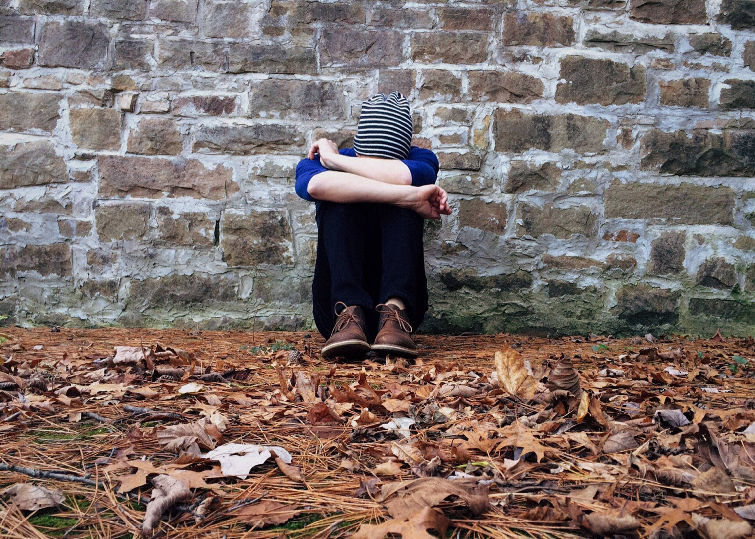mood 桌布 壁紙 — 一個人戴著條紋毛帽，蜷縮著靠在石牆上，雙手抱膝，周圍散落著秋天的落葉。