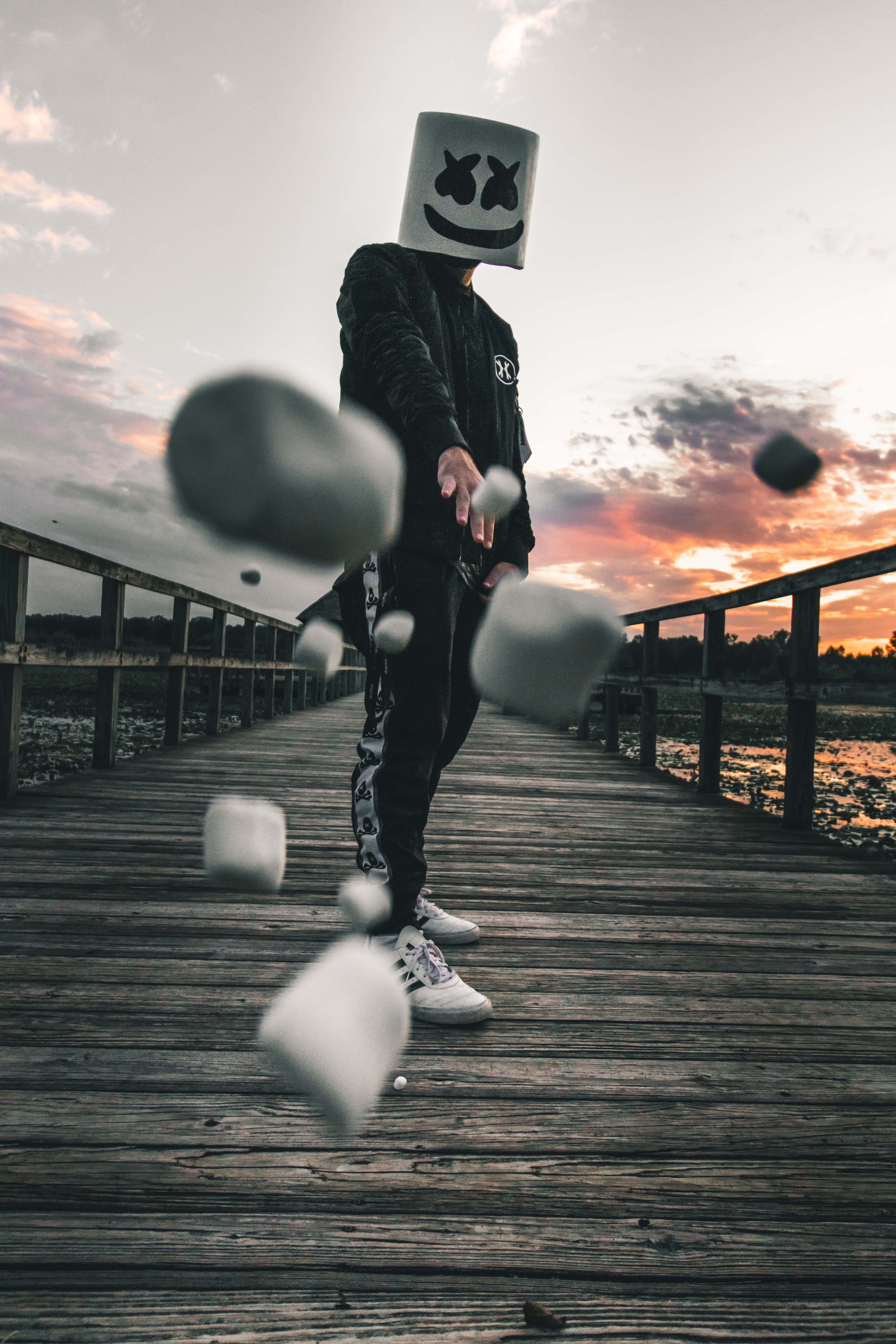Marshmello 動態圖 桌布 壁紙 — 一個人戴著棉花糖面具，在日落時分將漂浮的棉花糖扔在木製碼頭上。