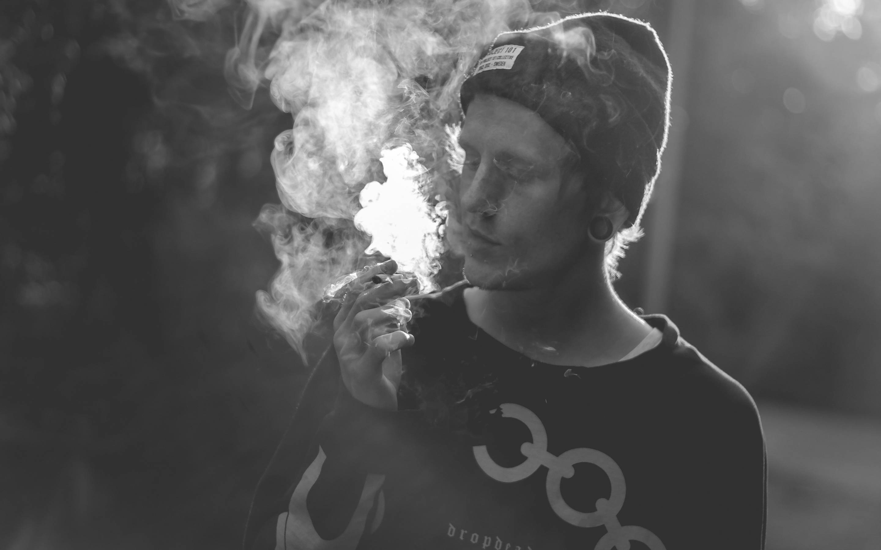 吸大麻 桌布 壁紙 — 一個人戴著毛帽，在模糊的戶外背景下，手持香煙呼出濃煙，以單色呈現。