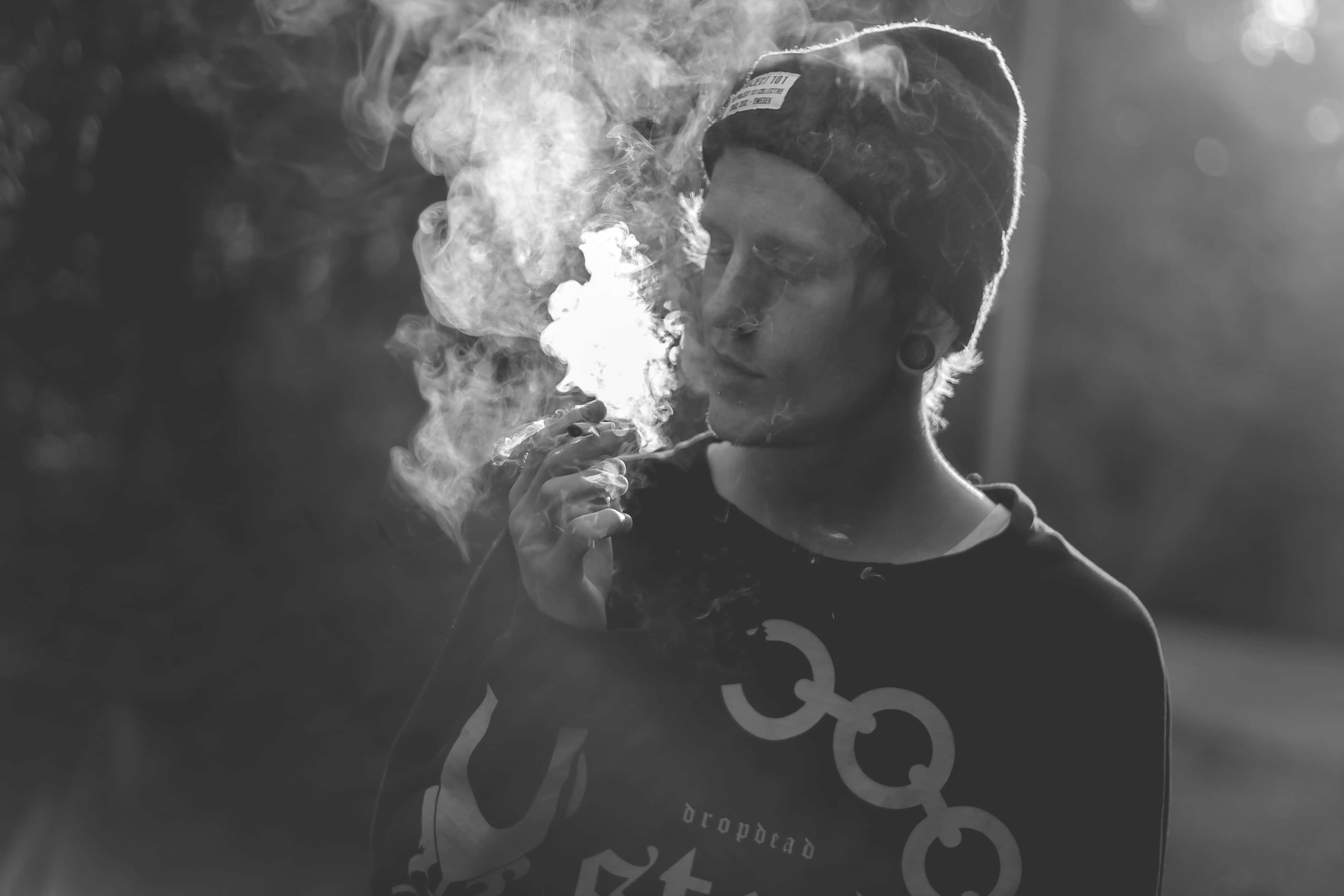 joint 桌布 壁紙 — 一個人戴著毛帽，在模糊的戶外背景下吸煙，呼出濃煙。