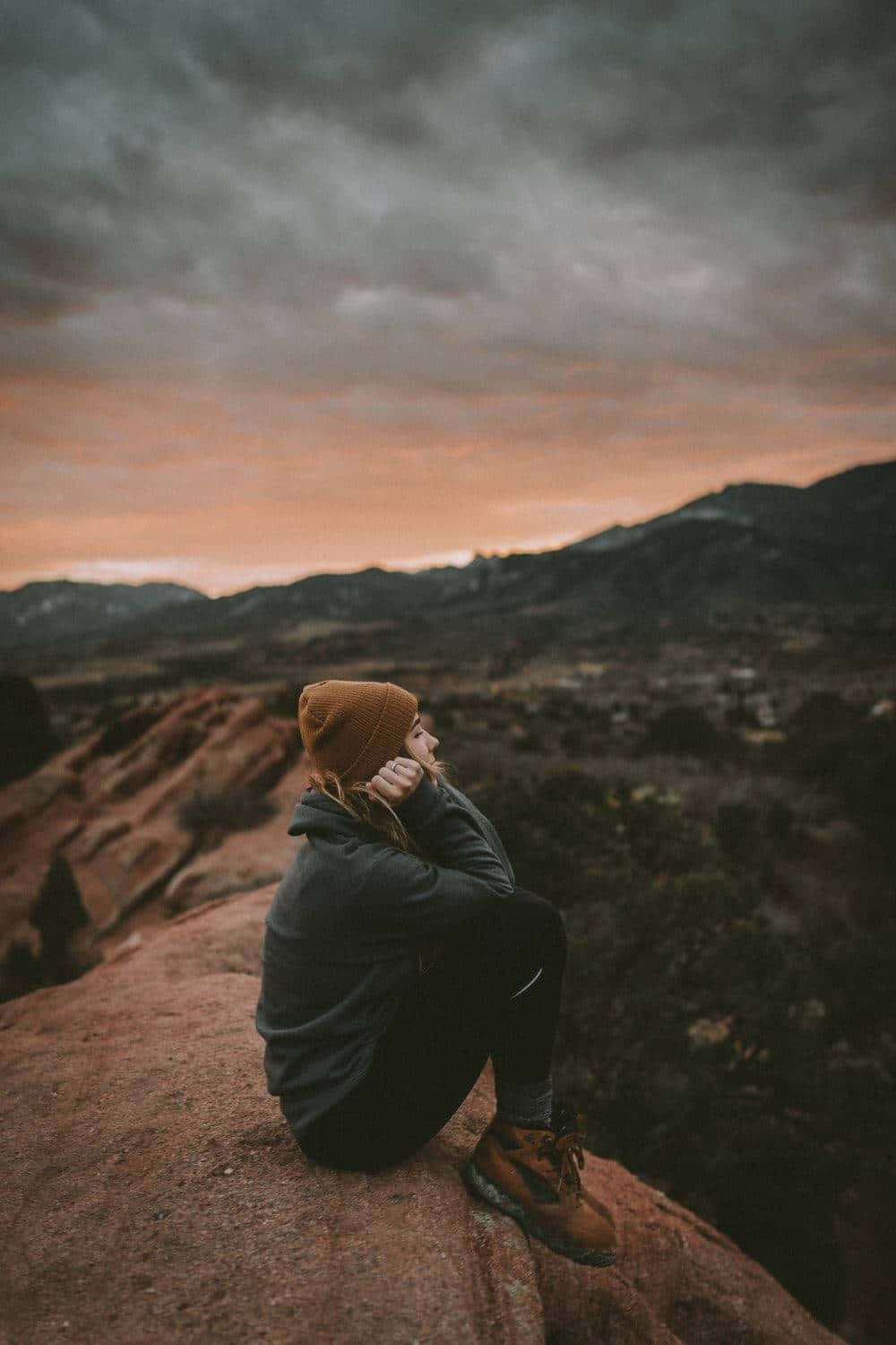 Alone 桌布 壁紙 — 一個人戴著毛帽獨自坐在紅色岩石懸崖上，在黃昏時分，凝視著在憂鬱的橙色和灰色天空下廣闊的山景。