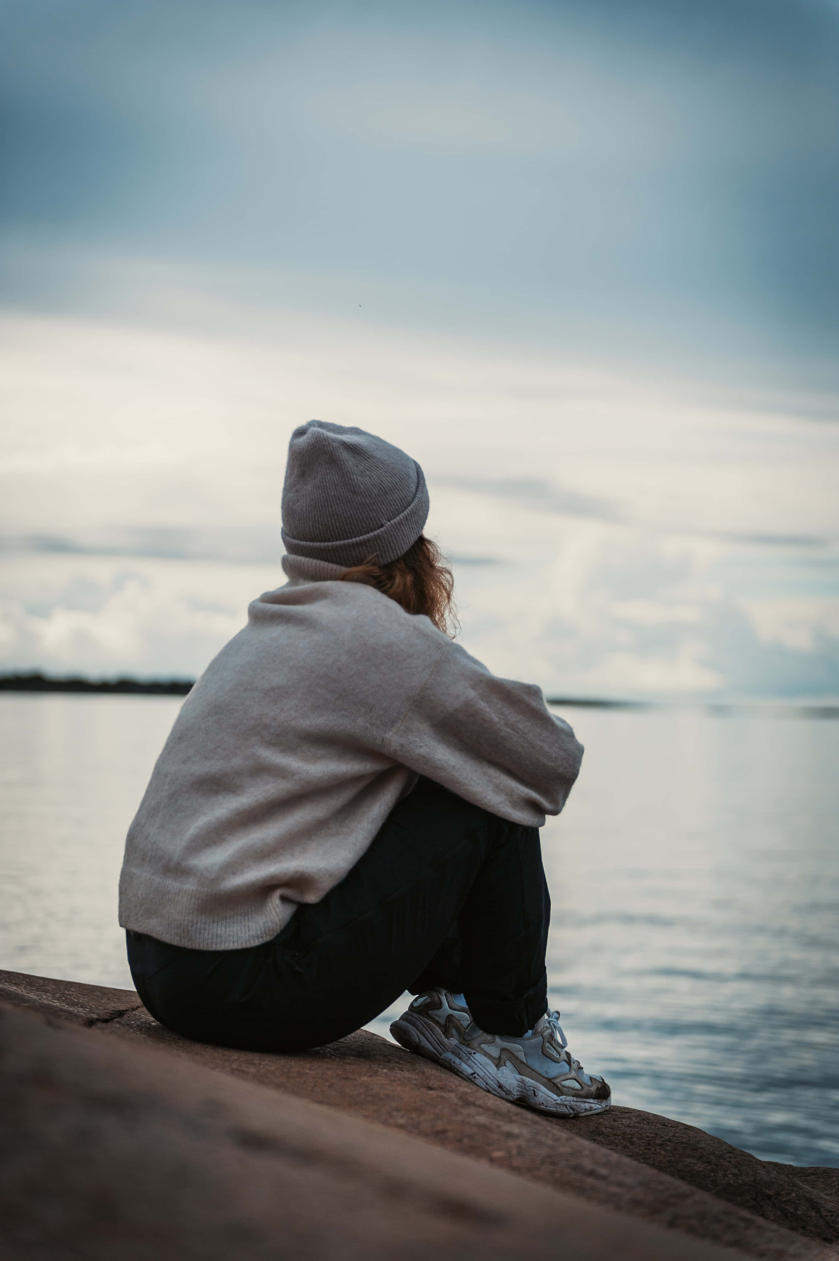 Single Girl 桌布 壁紙 — 一個人戴著灰色毛帽和連帽上衣，獨自坐在石頭邊緣，凝視著陰天下平靜的水面。