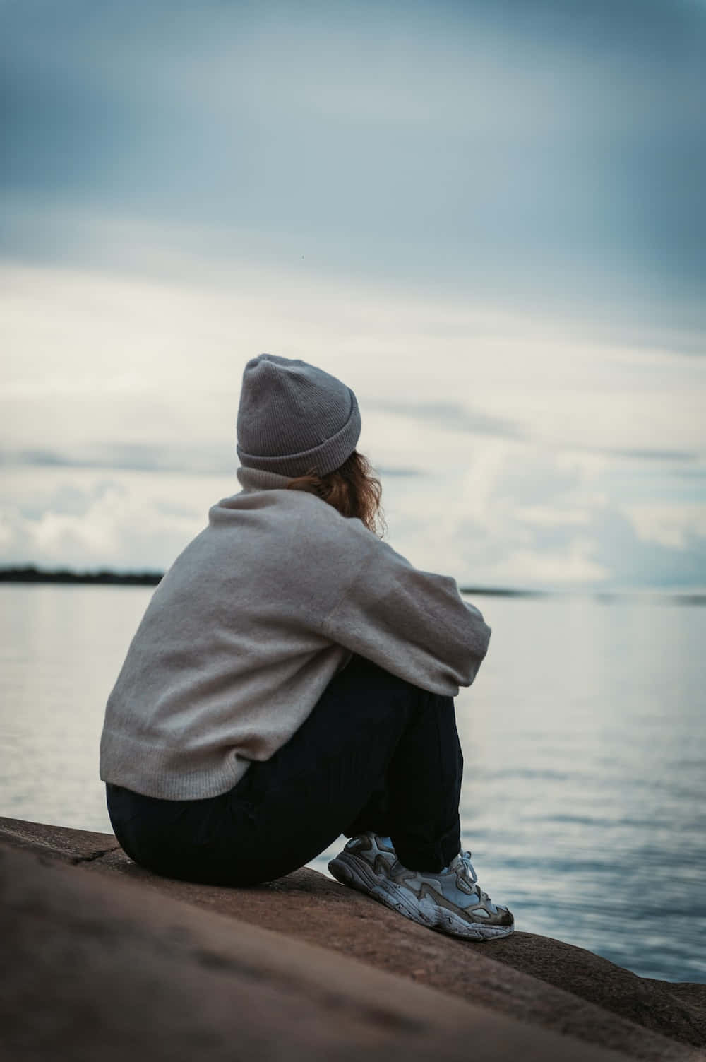 海洋 桌布 壁紙 — 一個人戴著灰色毛帽和連帽衫，獨自坐在石緣上，俯瞰著多雲天空下的平靜水面。
