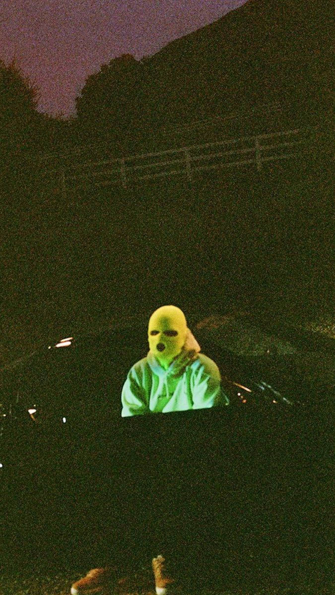 Travis Scott Aesthetic 桌布 壁紙 — 一個人戴著發光的黃色面具，坐在一輛黑暗的汽車裡，背景是紫色的黃昏天空、樹木和籬笆。