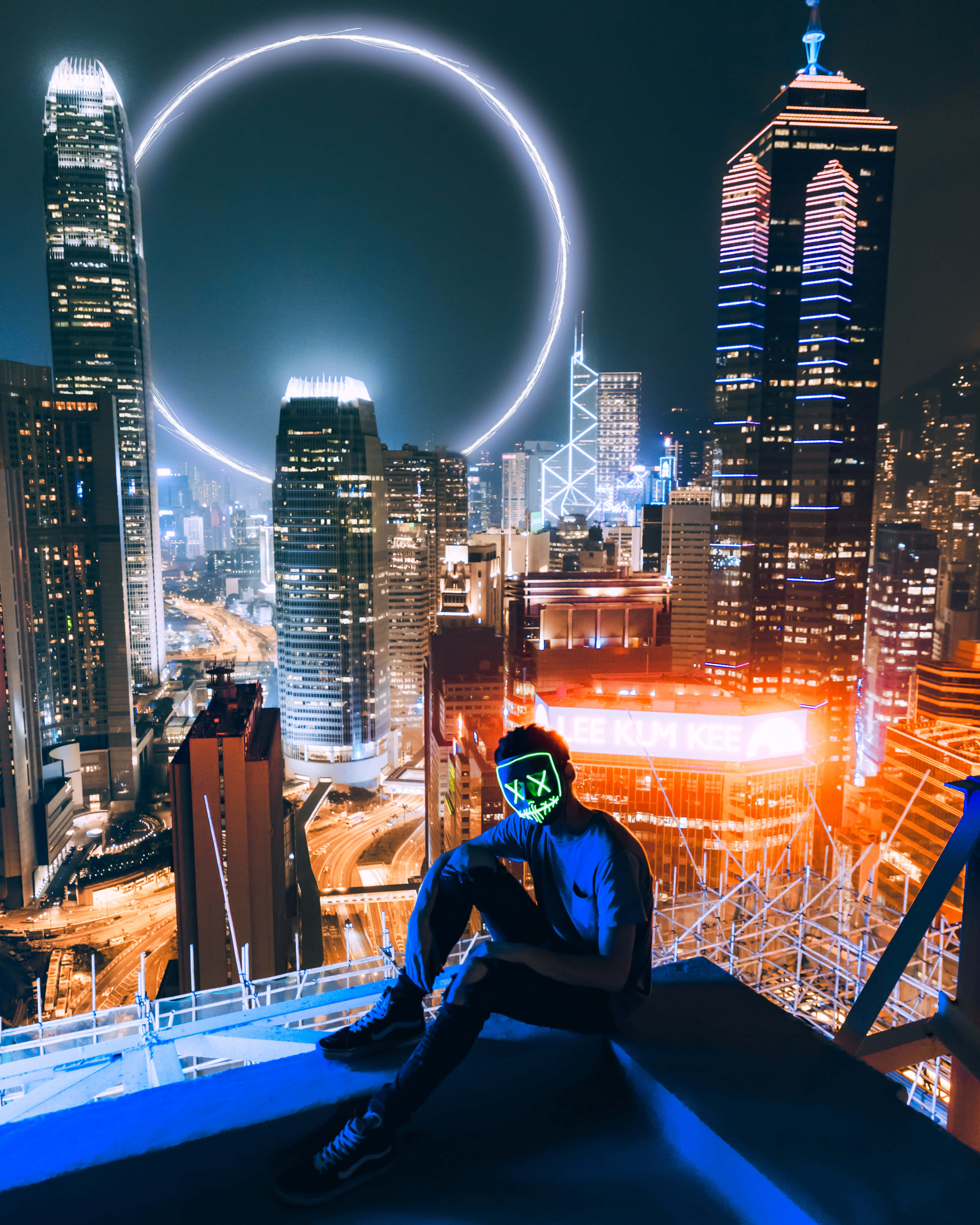 Rooftop 桌布 壁紙 — 一個人戴著發光綠色面具坐在摩天大樓邊緣，俯瞰著霓虹燈閃爍的城市景觀，夜空中有發光的圓形光環。
