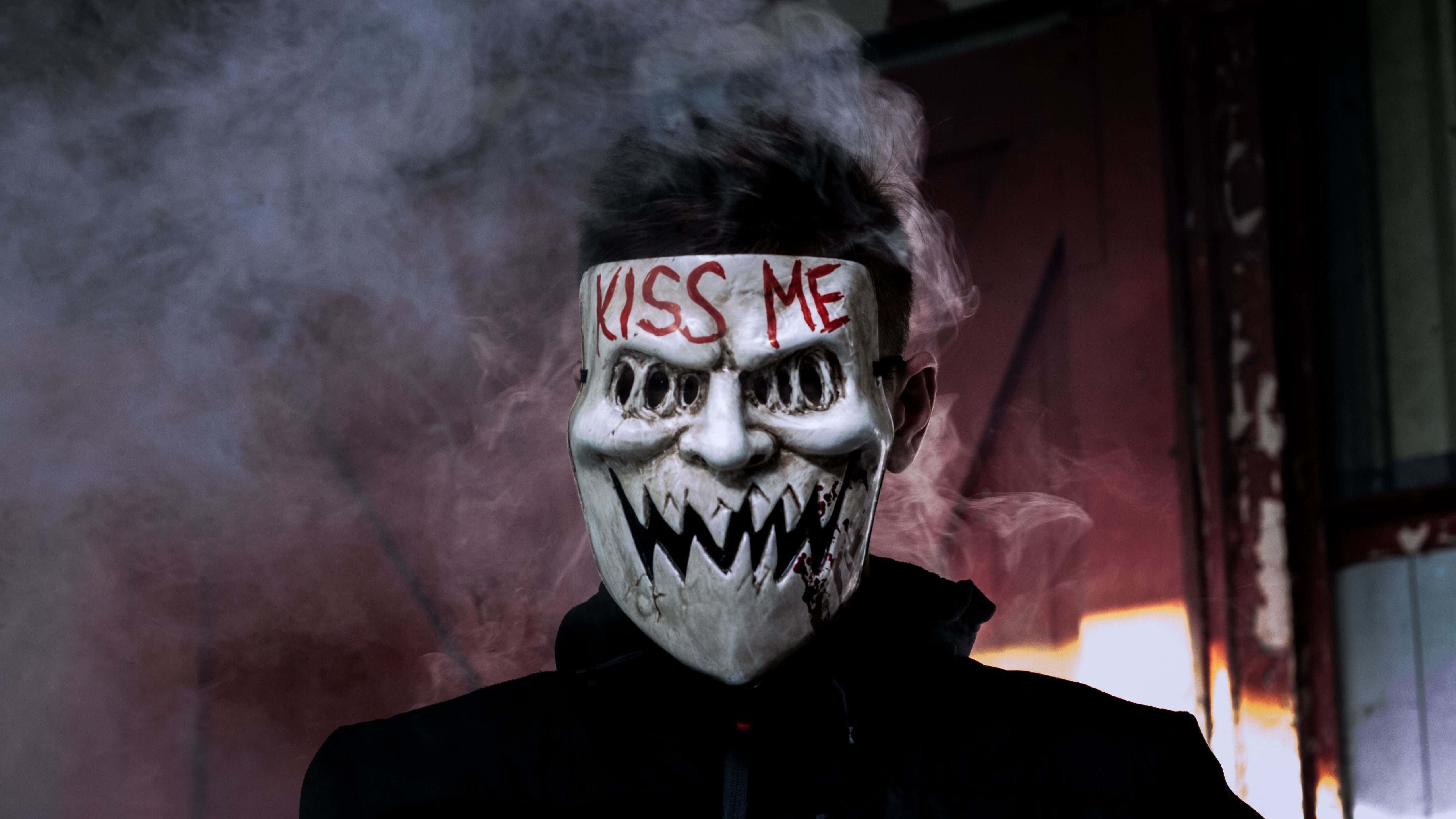 Halloween Masks 桌布 壁紙 — 一個人戴著白色恐怖面具，上面有紅色「KISS ME」文字和鋸齒狀牙齒，站在煙霧瀰漫的昏暗光線中，靠近燃燒的門口。