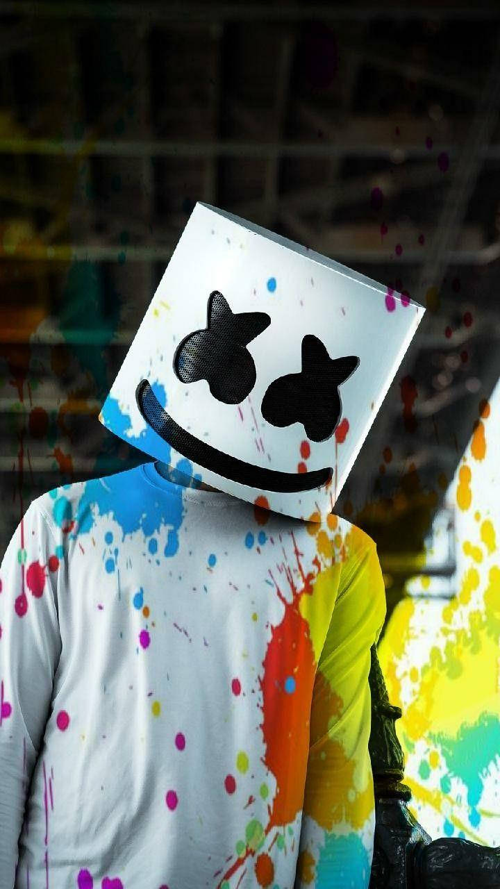 marshmello iPhone 桌布 壁紙 — 一個人戴著白色方形面具，面具上有星形眼睛和微笑，身上佈滿彩色油漆噴濺，背景是模糊的工業環境。