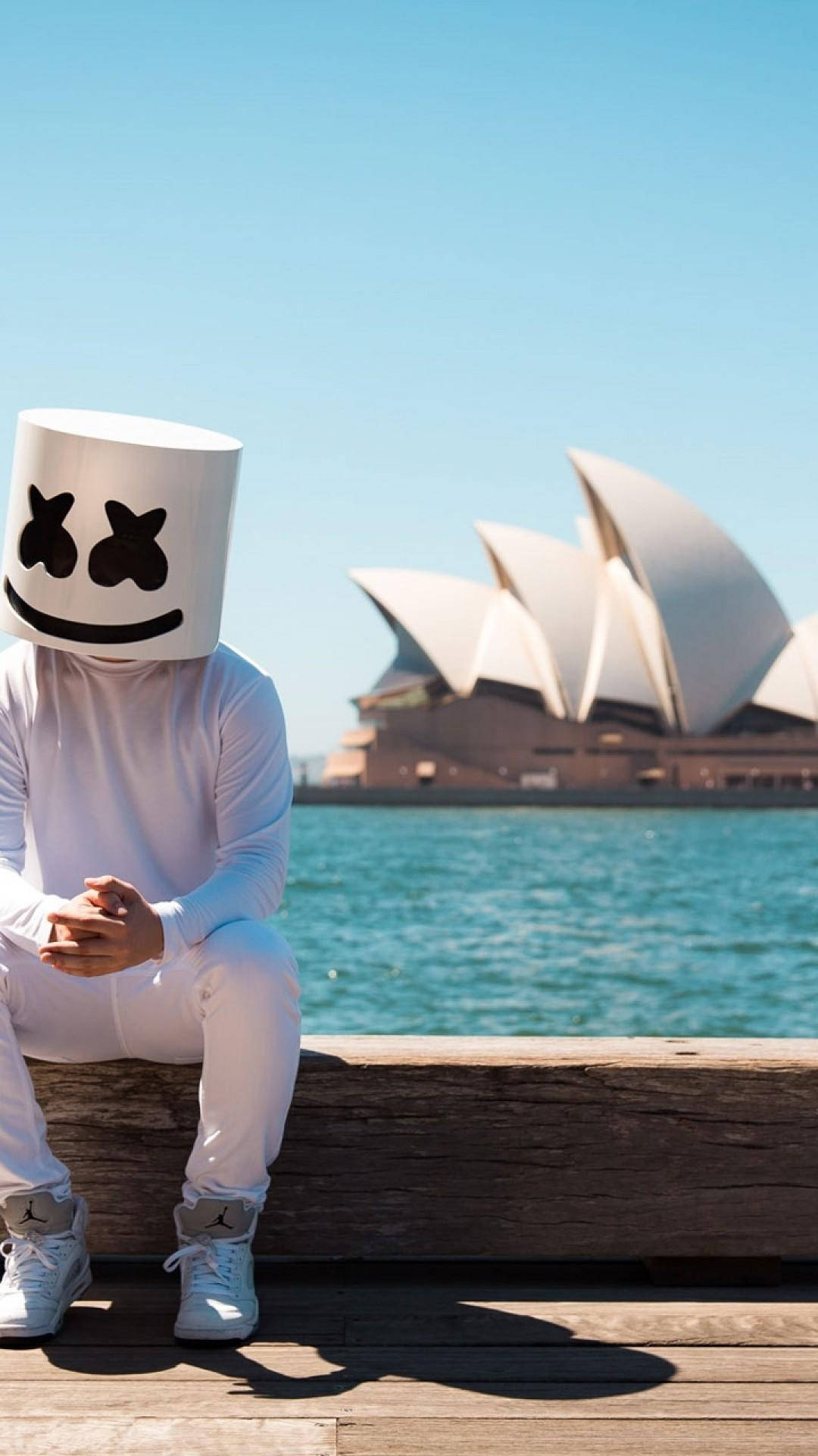 marshmello iPhone 桌布 壁紙 — 一個人戴著白色棉花糖面具和穿著白色棉花糖服裝，坐在木製碼頭上，背景是雪梨歌劇院和藍色海水，天空晴朗。