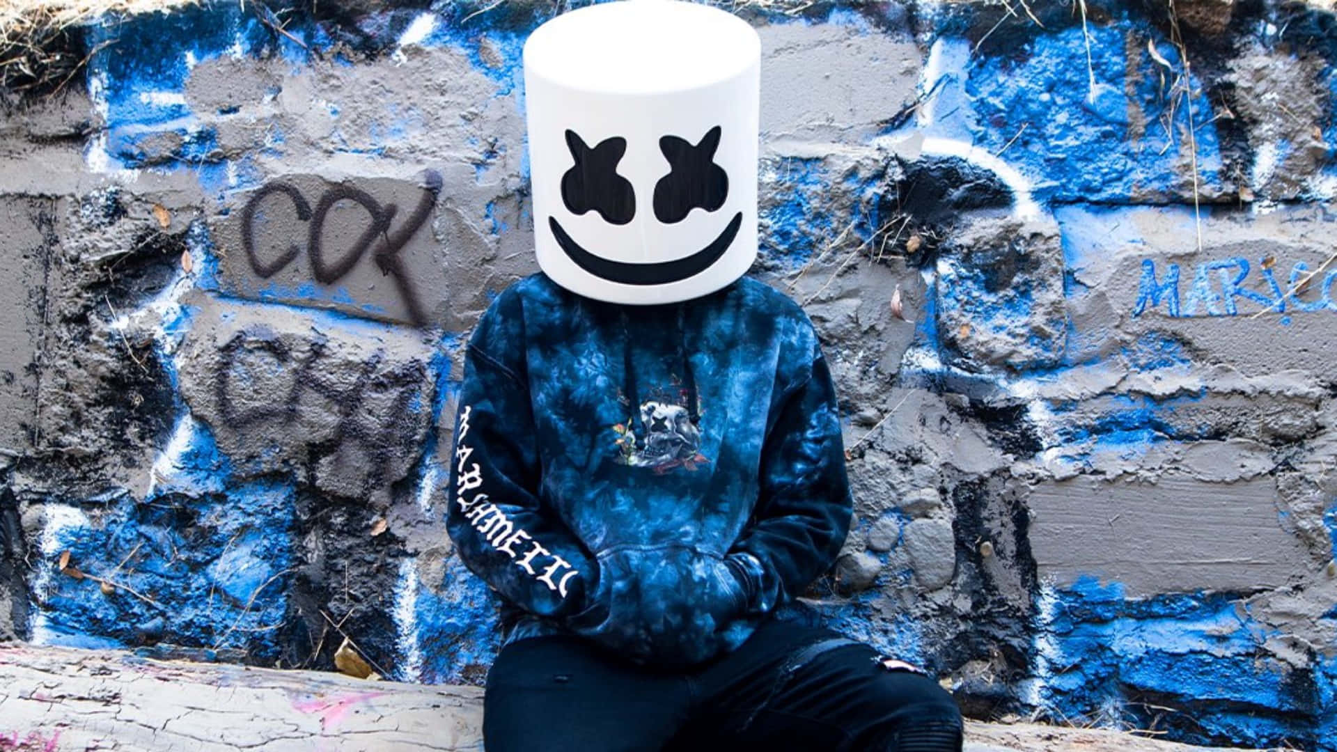 Marshmello 動態圖 桌布 壁紙 — 一個人戴著白色棉花糖頭盔、身穿藍色紮染連帽衫，靠著佈滿塗鴉的磚牆坐著。