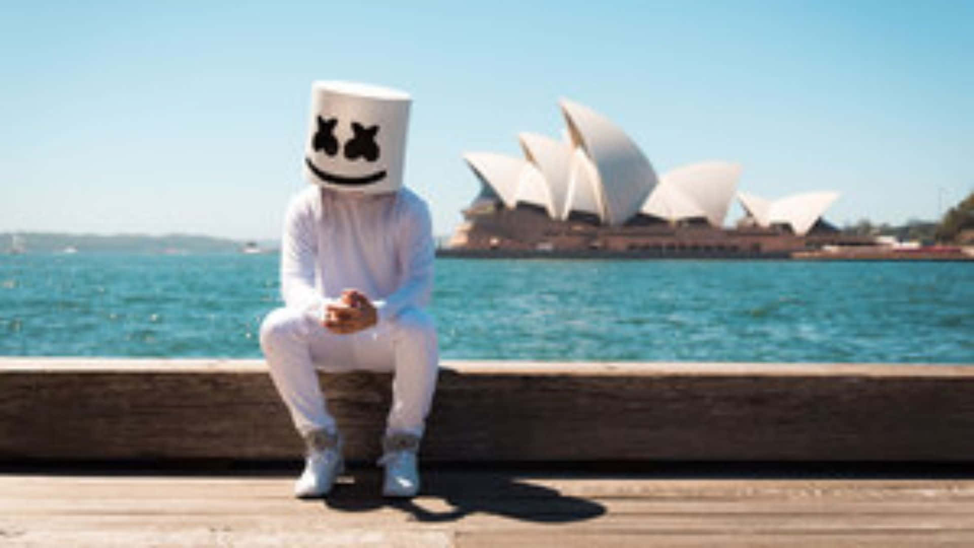 Marshmello 動態圖 桌布 壁紙 — 一個人戴著白色棉花糖頭盔坐在木碼頭上，背景是雪梨歌劇院，天空清澈湛藍。