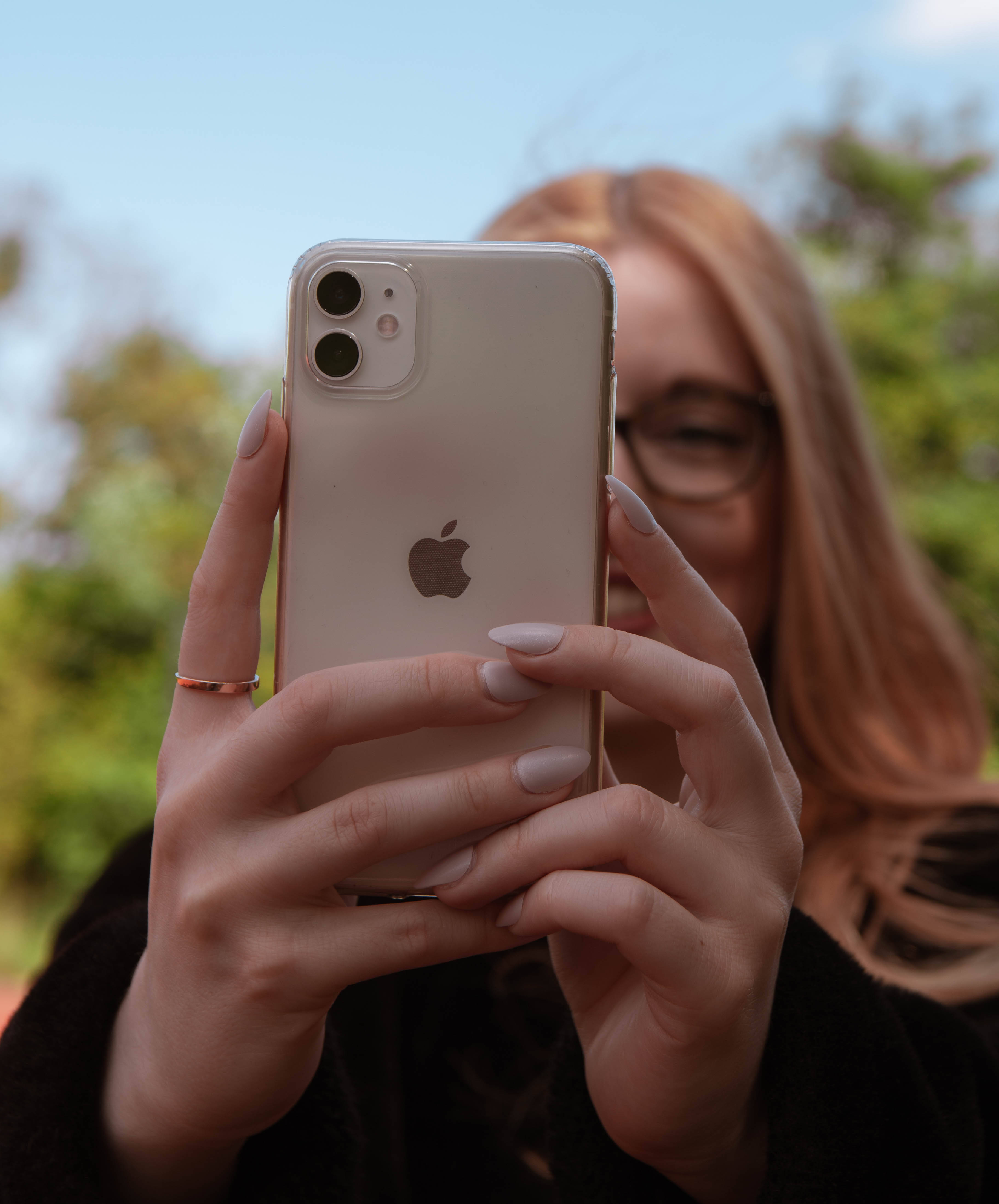 iPhone 11 桌布 壁紙 — 一個人戴著眼鏡，淺棕色頭髮，拿著金色iPhone 11在模糊的綠色戶外背景前自拍。