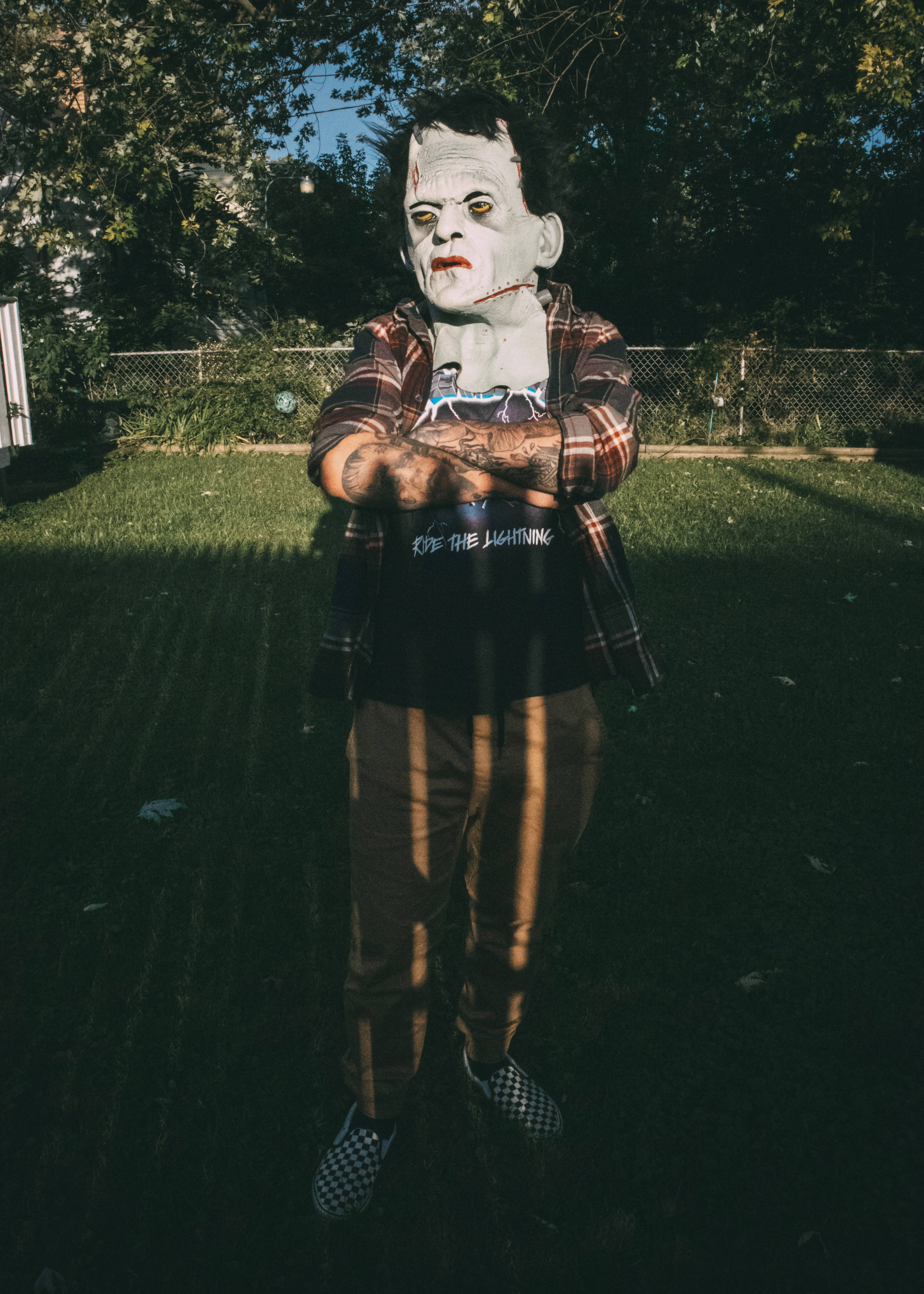 Vintage Halloween 桌布 壁紙 — 一個人戴著科學怪人面具，雙臂交叉，露出刺青，站在陽光普照的後院。