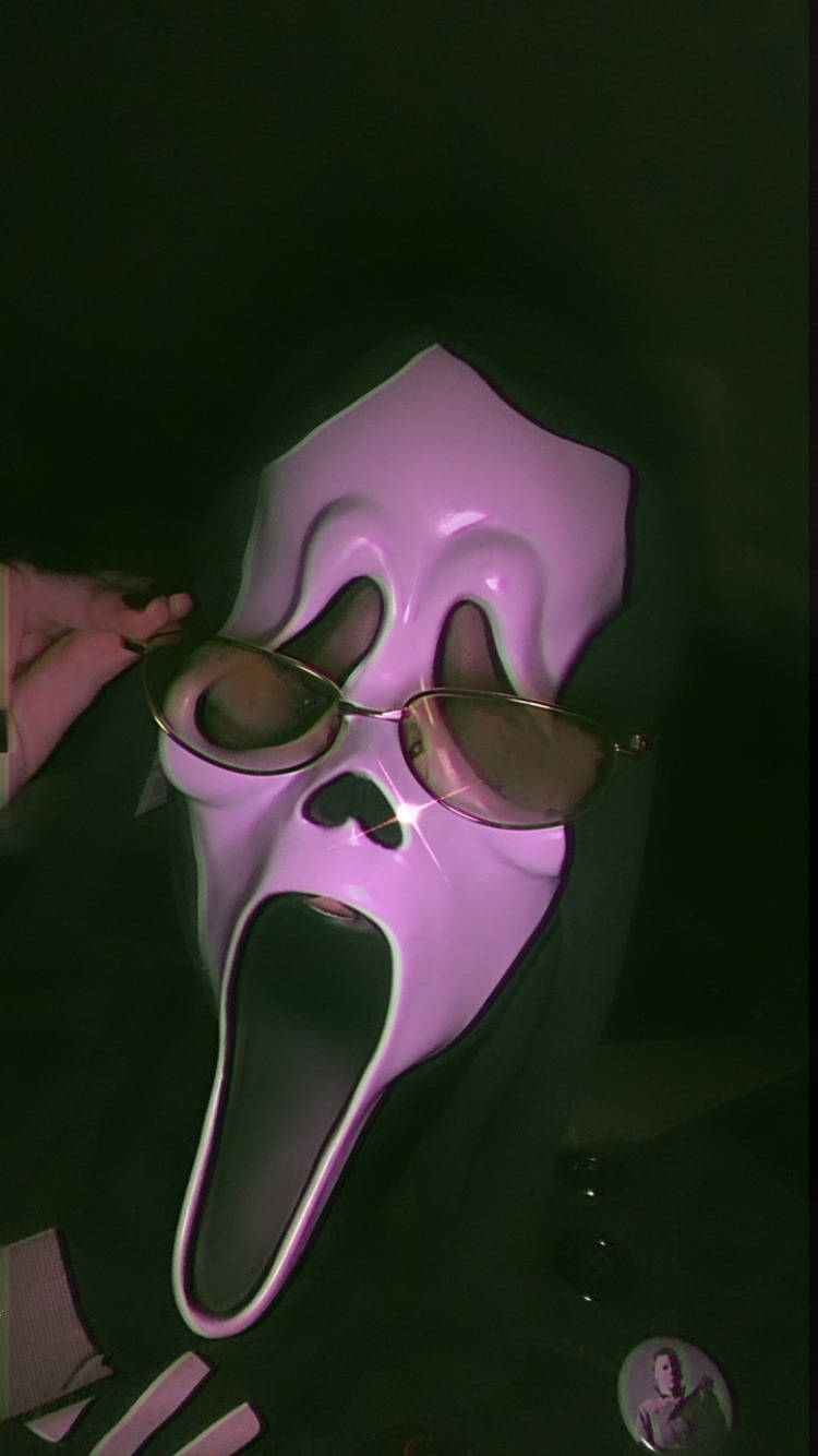 吶喊 桌布 壁紙 — 一個人戴著粉紅色 Ghostface 面具，在昏暗的綠色燈光下戴著金色框太陽眼鏡。