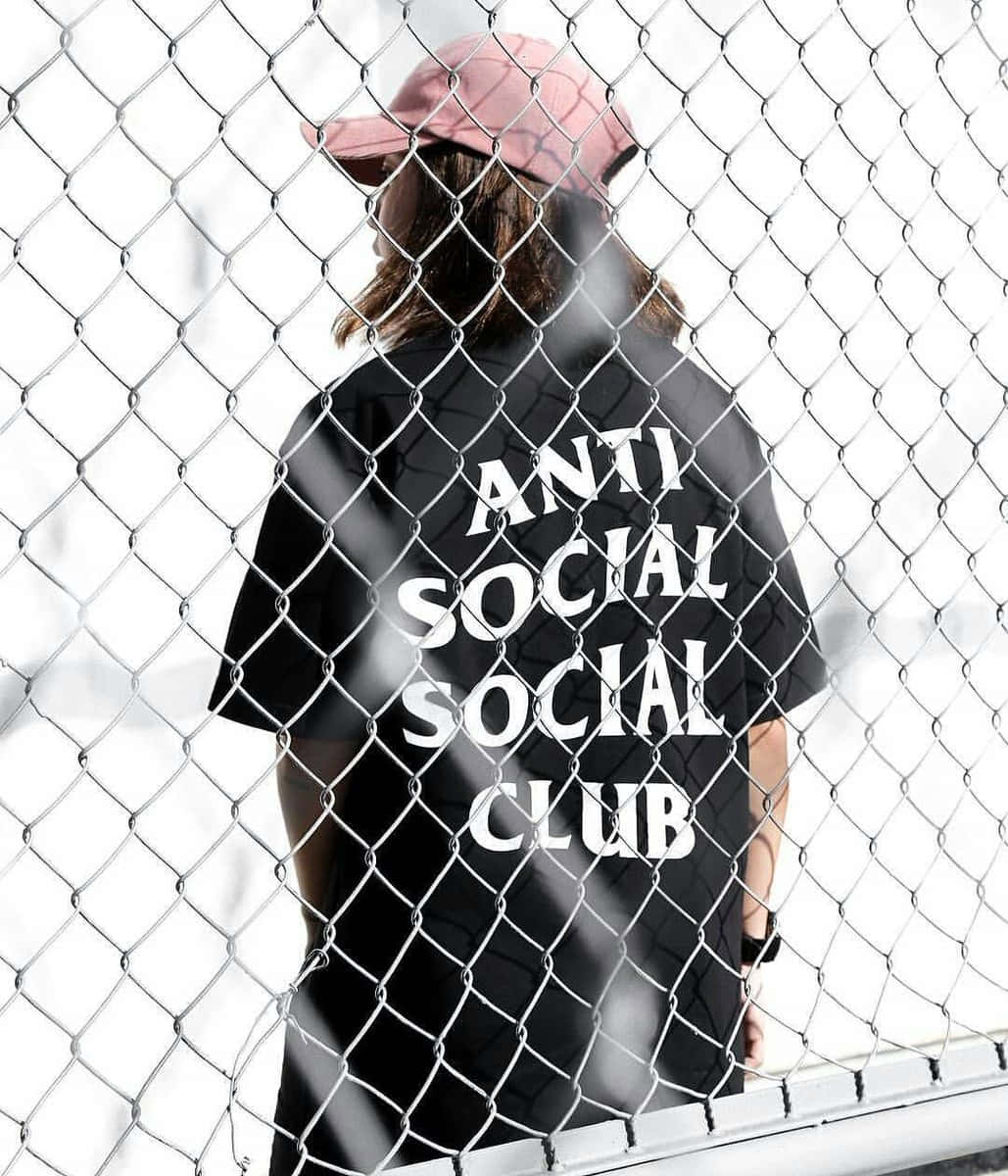 Anti Social Club iPhone 桌布 壁紙 — 一個人戴著粉紅色帽子，穿著黑色'ANTI SOCIAL SOCIAL CLUB'襯衫，站在鐵絲網後面，從背後看。