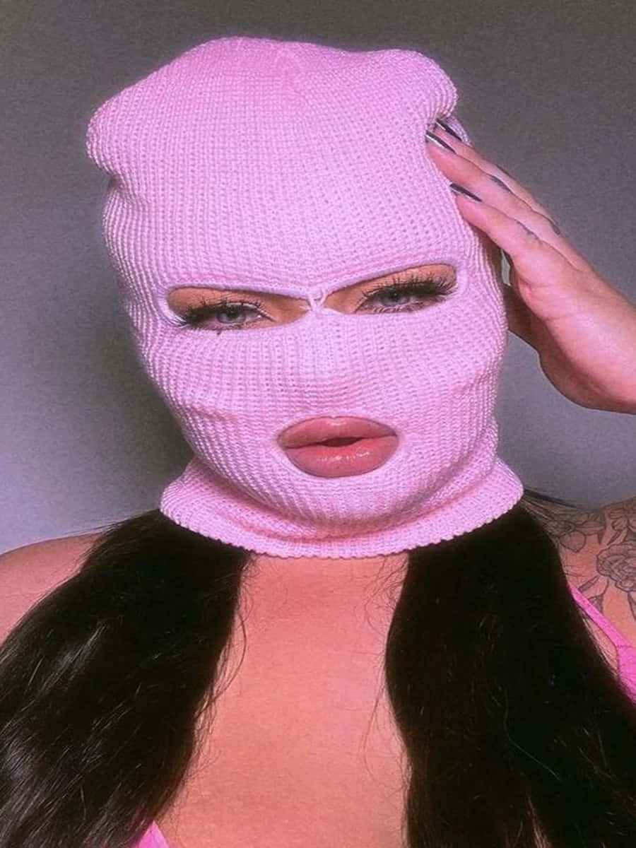 Ski Mask Girl 桌布 壁紙 — 一個人戴著粉紅色針織巴拉克拉法帽，只露出眼睛和嘴唇，手觸摸頭部，背景為純灰色。
