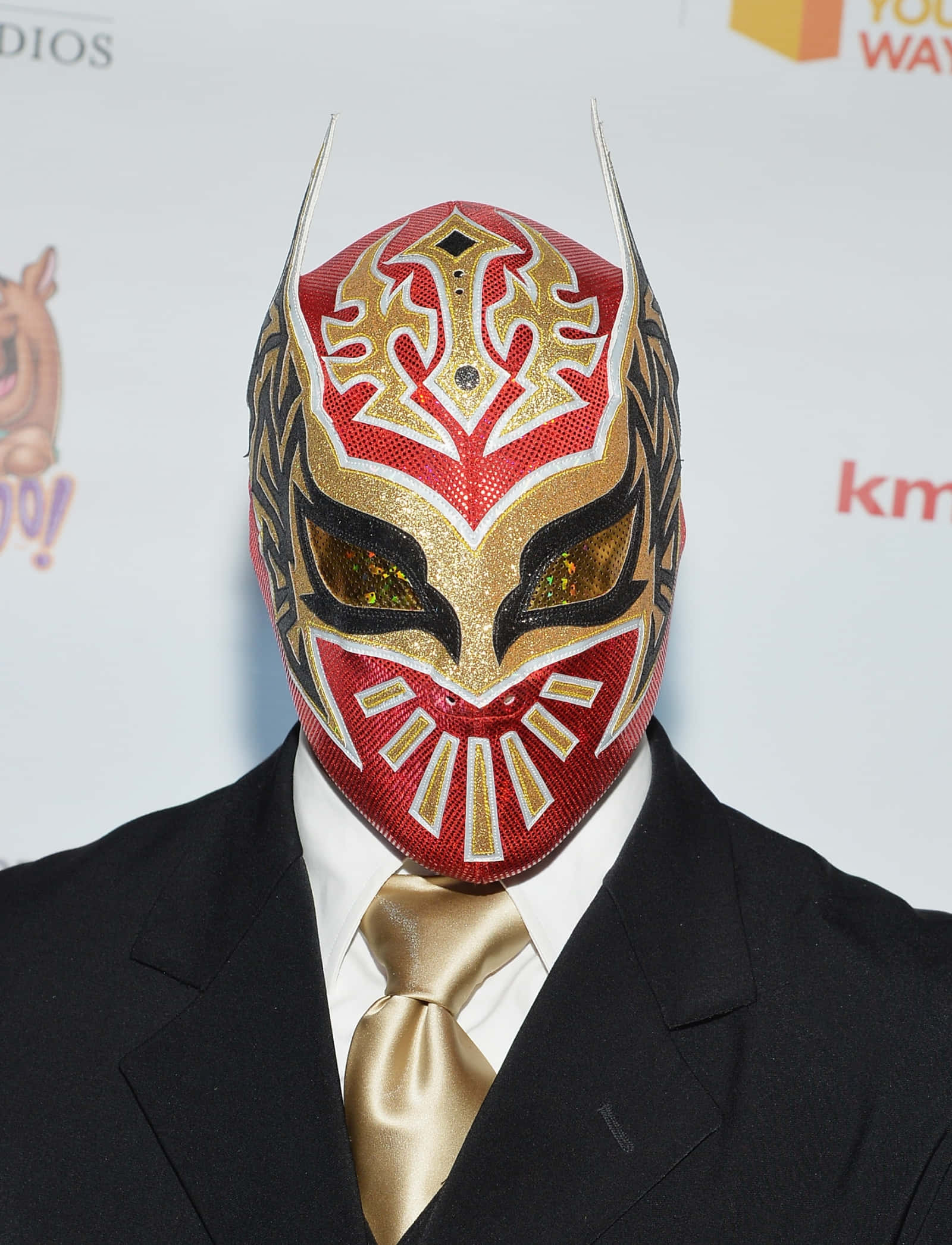 WWE 桌布 壁紙 — 一個人戴著紅、金、黑色華麗自由摔角面具，穿著正式黑色西裝和金色領帶擺姿勢。