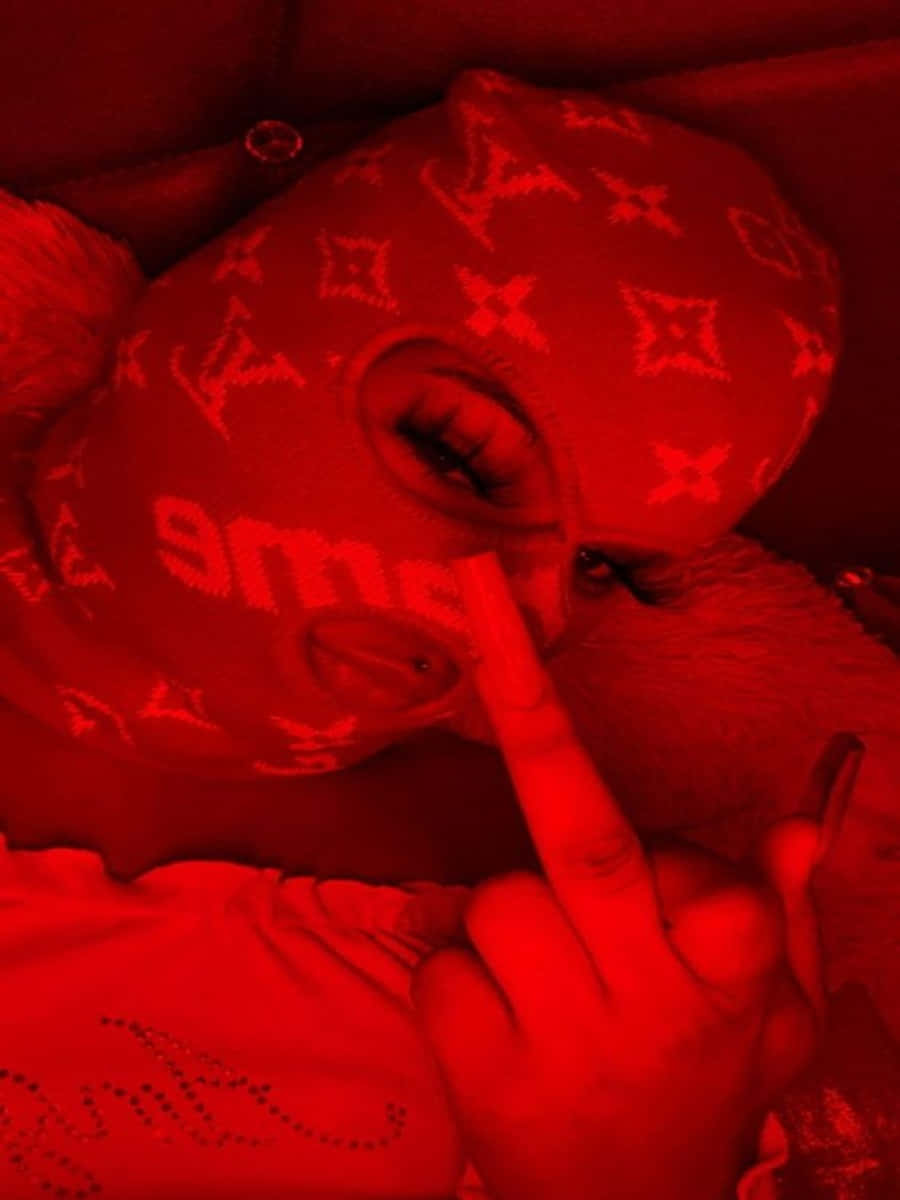 Ski Mask Girl 桌布 壁紙 — 一個人戴著紅色 Louis Vuitton 圖案面具，露出一隻眼睛，手指放在嘴唇上，沐浴在單色紅光中。