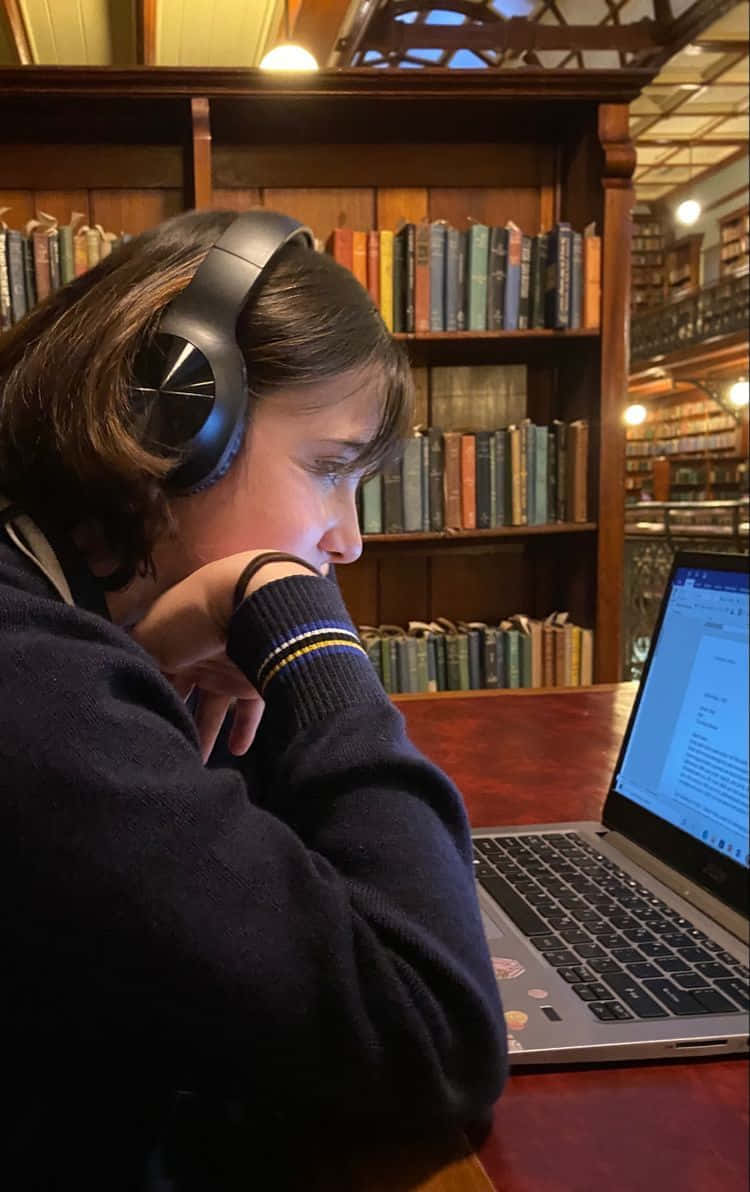Studying 桌布 壁紙 — 一個人戴著耳機，在燈光溫暖、有高聳木書架的圖書館裡專注地使用筆記型電腦。