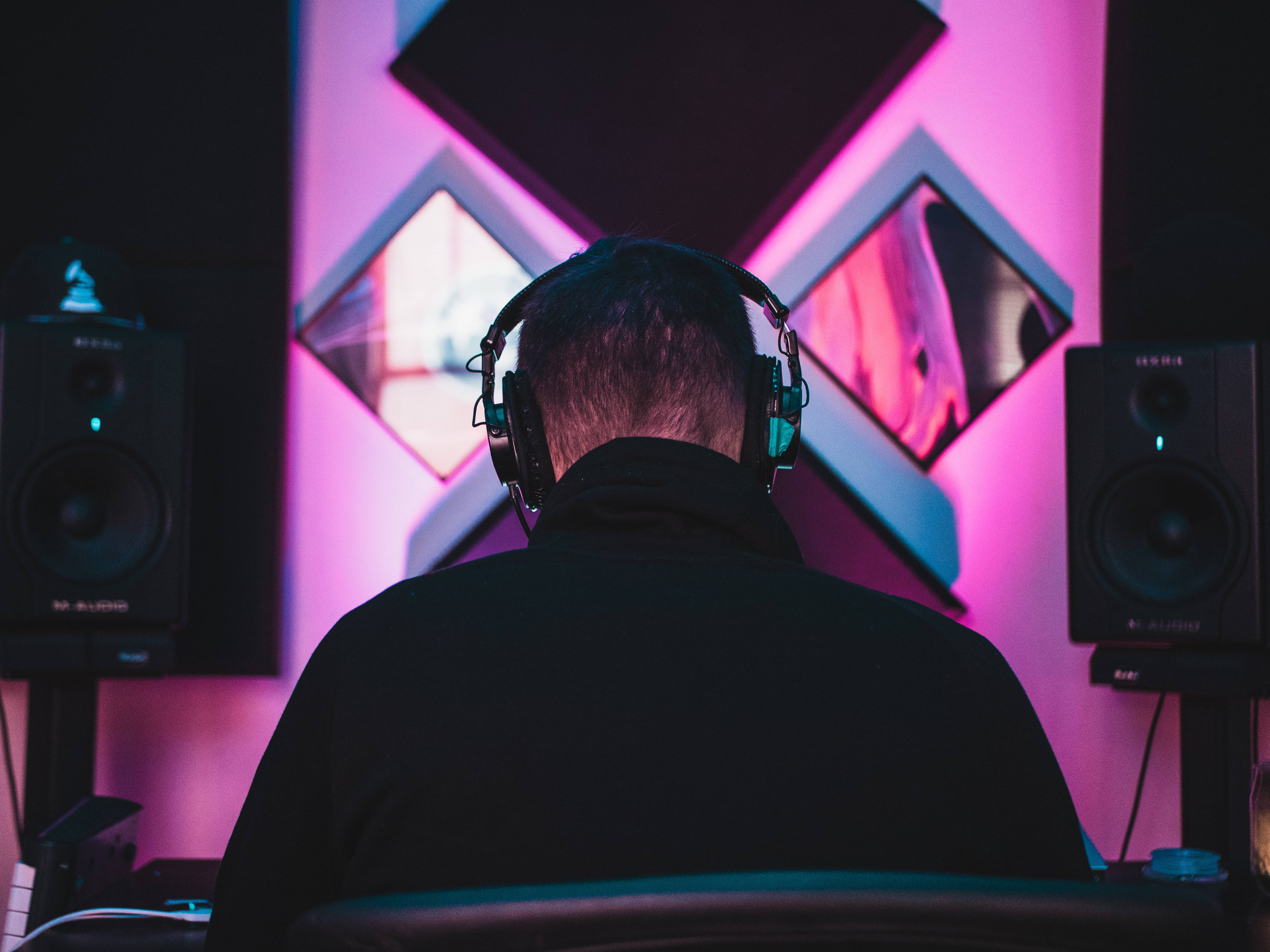 DJ 桌布 壁紙 — 一個人戴著耳機坐在光線昏暗的錄音室中，被幾何牆板和揚聲器發出的鮮豔粉紅和藍色霓虹燈光所籠罩。