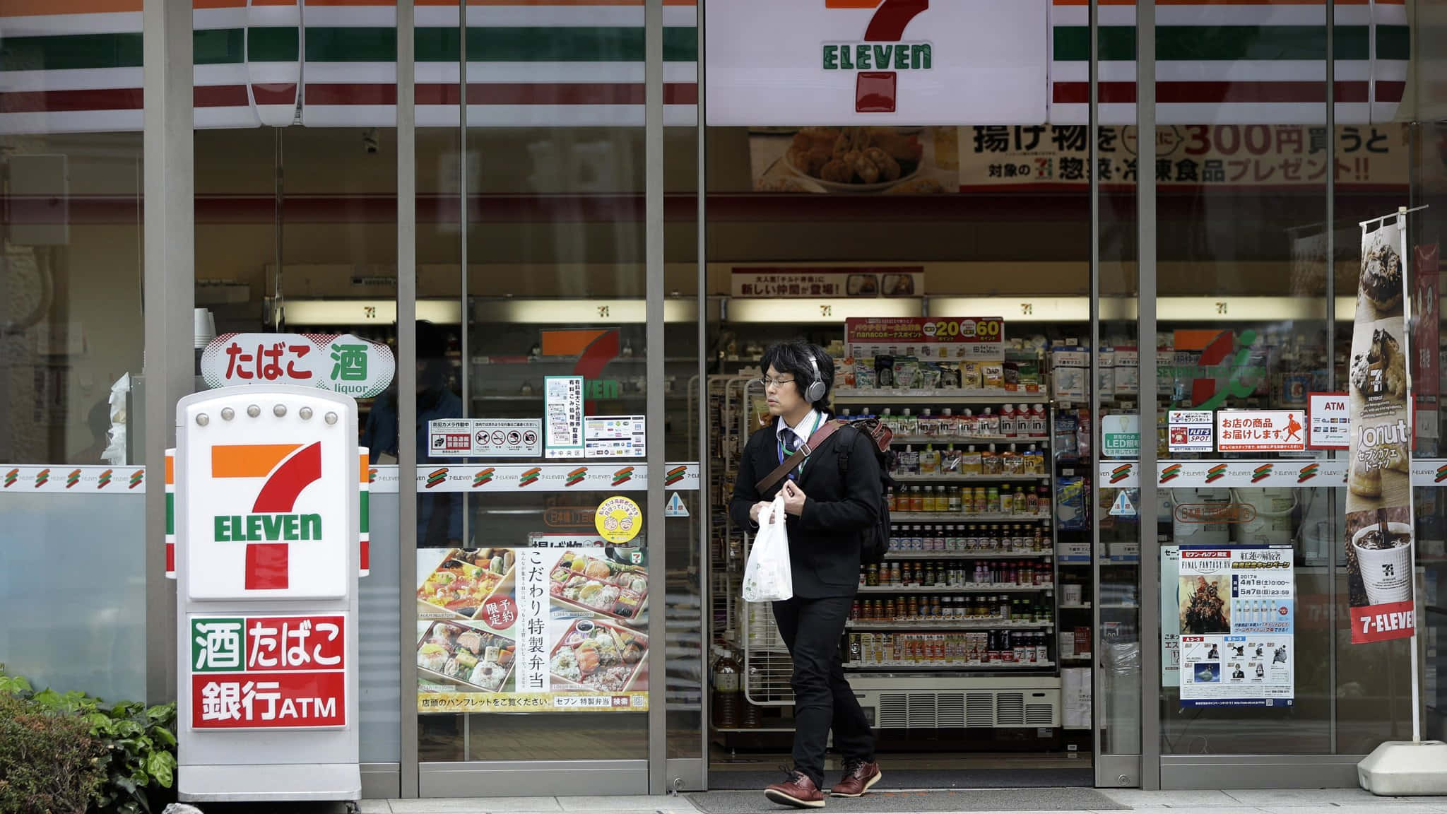 7-ELEVEN 桌布 壁紙 — 一個人戴著耳機走過一間燈火通明的7-Eleven店鋪，店鋪有日文招牌和玻璃門反射街道。