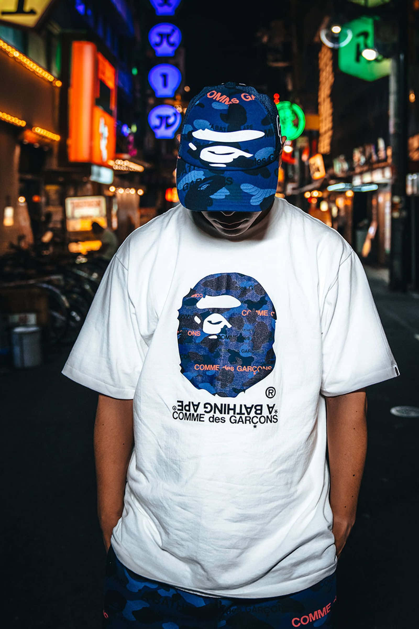Bape 動態 桌布 壁紙 — 一個人戴著藍色迷彩帽，穿著白色A Bathing Ape T恤，夜晚站在霓虹燈閃爍的東京街道上。