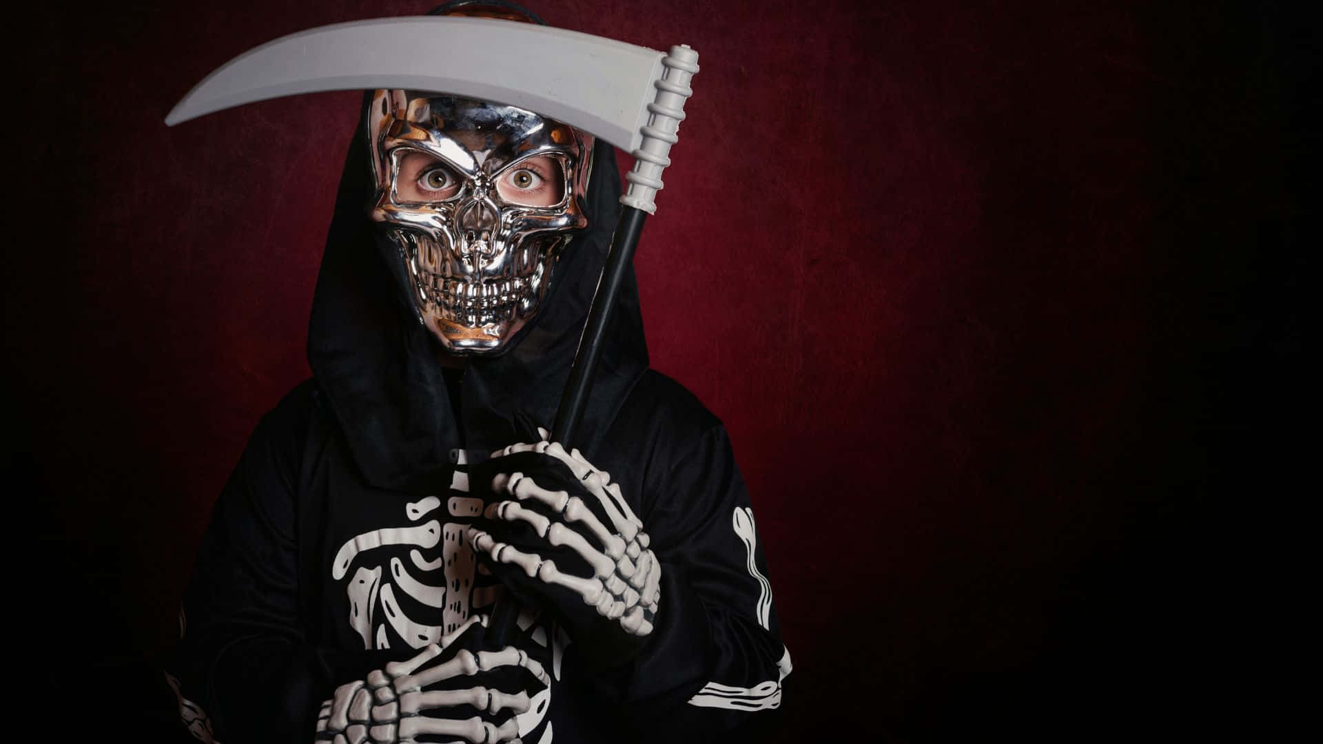 Skeleton Costumes 桌布 壁紙 — 一個人戴著金屬骷髏面具和黑色連帽服裝，手持鐮刀，背景是深紅色。