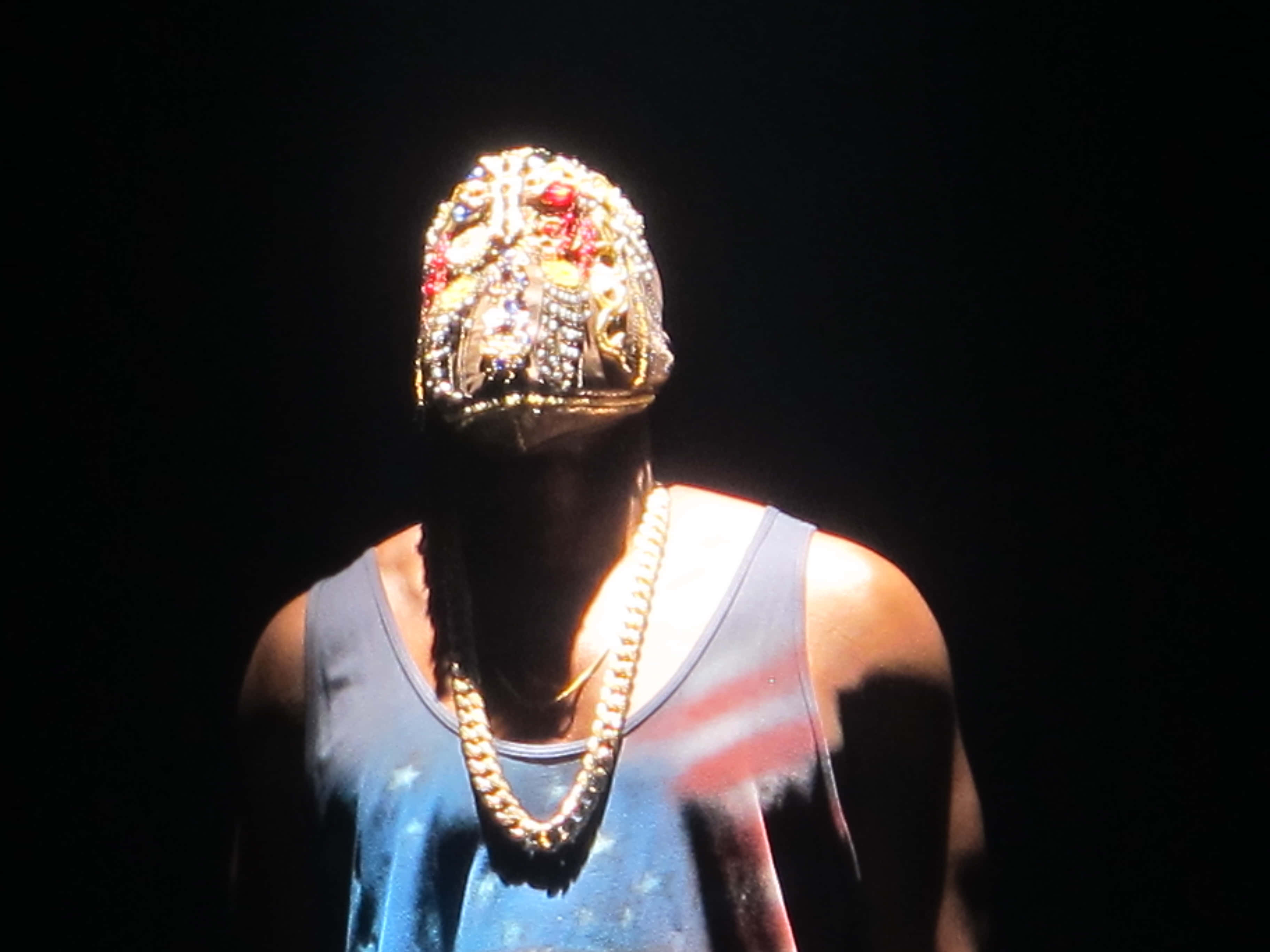 Kanye West Yeezus 桌布 壁紙 — 一個人戴著鑲有寶石的面具和粗鏈條，在黑色背景下的戲劇性聚光燈下。