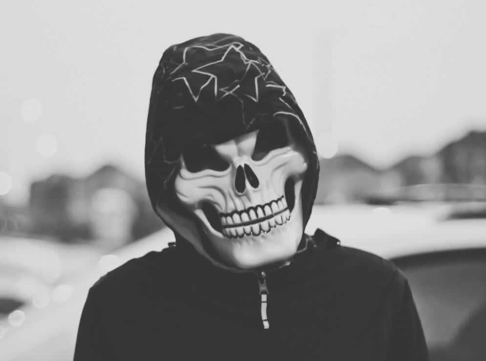Halloween Masks 桌布 壁紙 — 一個人戴著骷髏面具和連帽外套，身處單色調的城市環境中，背景燈光模糊。