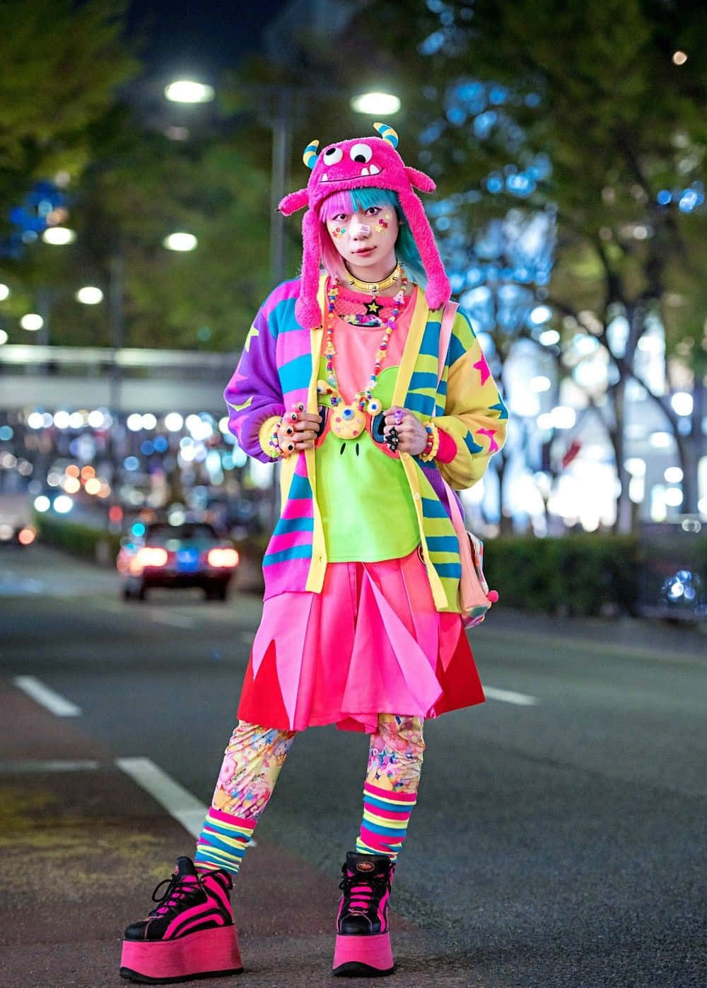 Harajuku Fashion 桌布 壁紙 — 一個人戴著鮮豔的粉紅色怪物帽子和色彩繽紛的街頭服飾，自信地站在夜晚的城市街道上，背後有模糊的燈光。