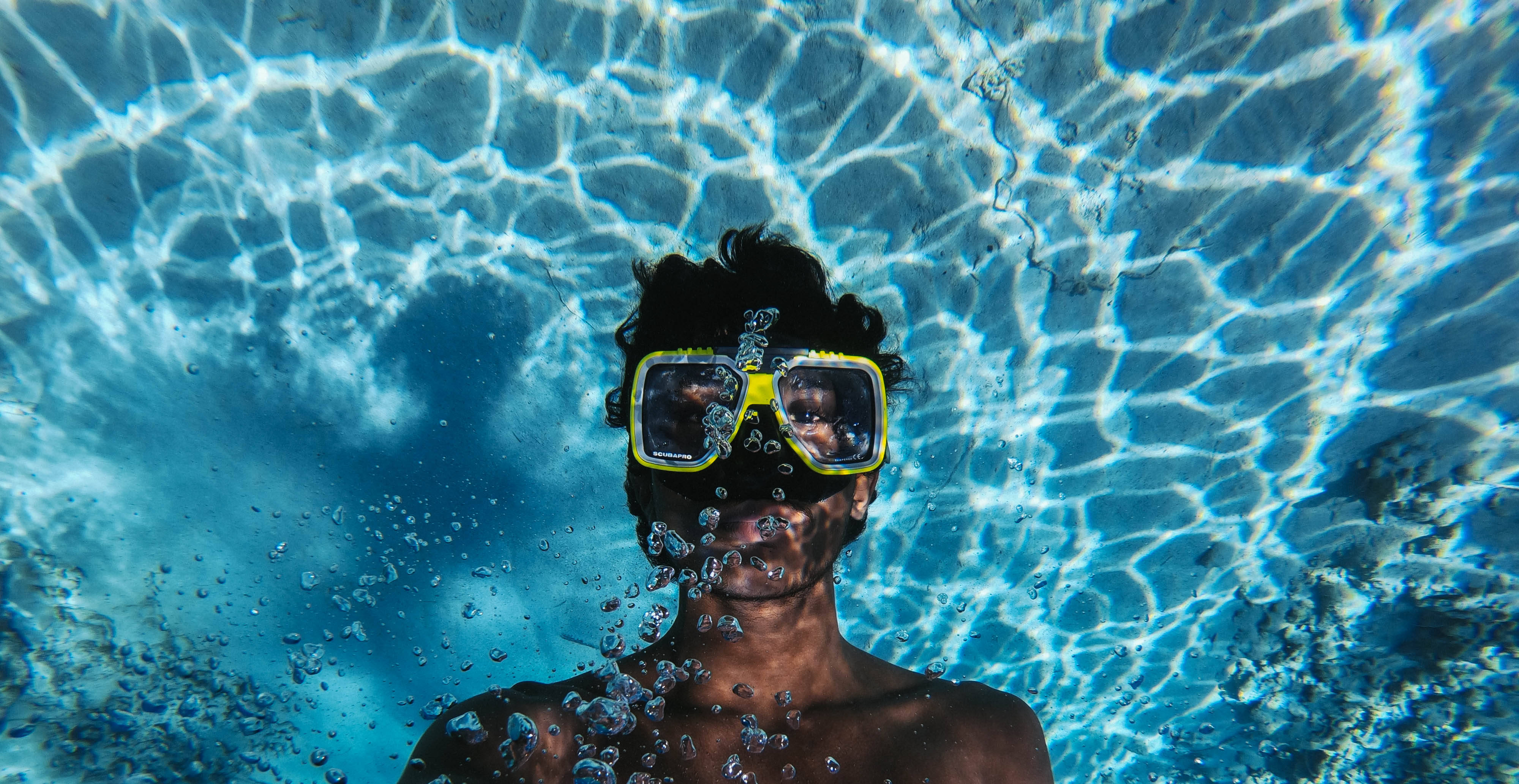 Snorkeling 桌布 壁紙 — 一個人戴著黃色泳鏡從水下向上看，陽光在藍色泳池表面泛起漣漪。