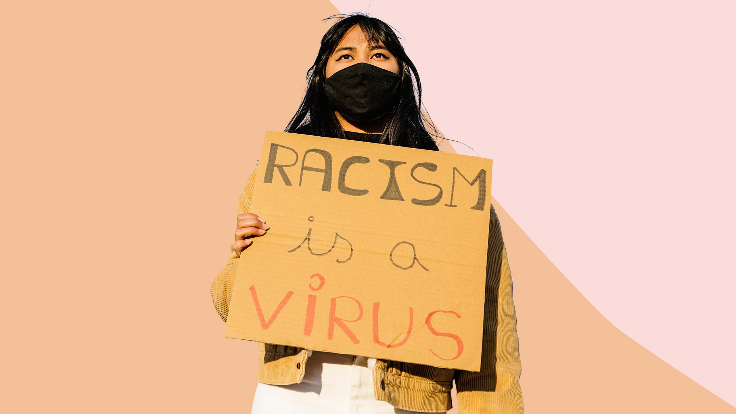 Woman 桌布 壁紙 — 一個人戴著黑色口罩，舉著一個紙板標語，上面寫著「RACISM is a VIRUS」，背景是柔和的蜜桃色和粉紅色。