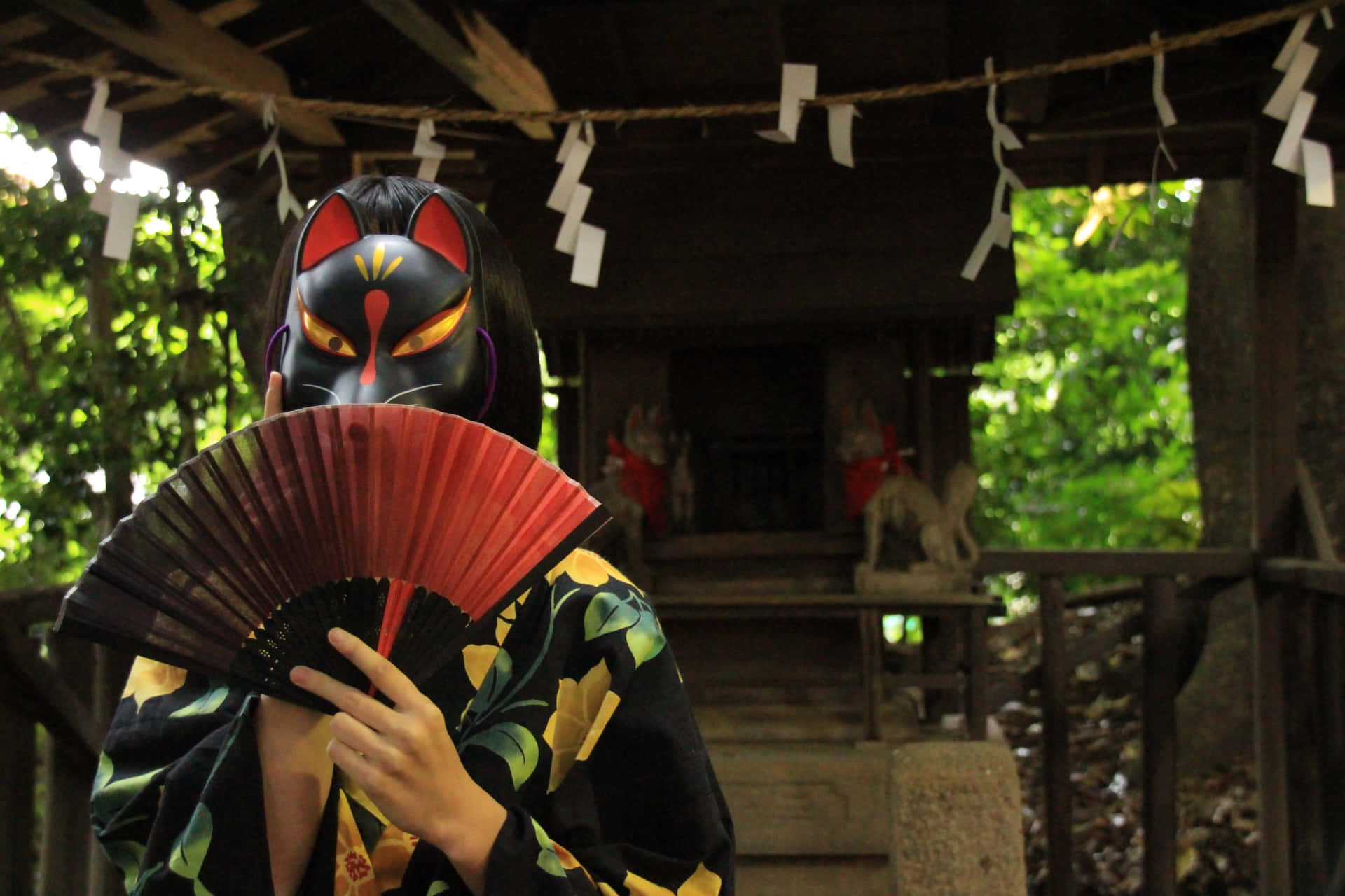 Tengu 桌布 壁紙 — 一個人戴著黑色狐狸面具、穿著花卉和服，在木造神社拿著紅色扇子，周圍是茂密的綠葉。