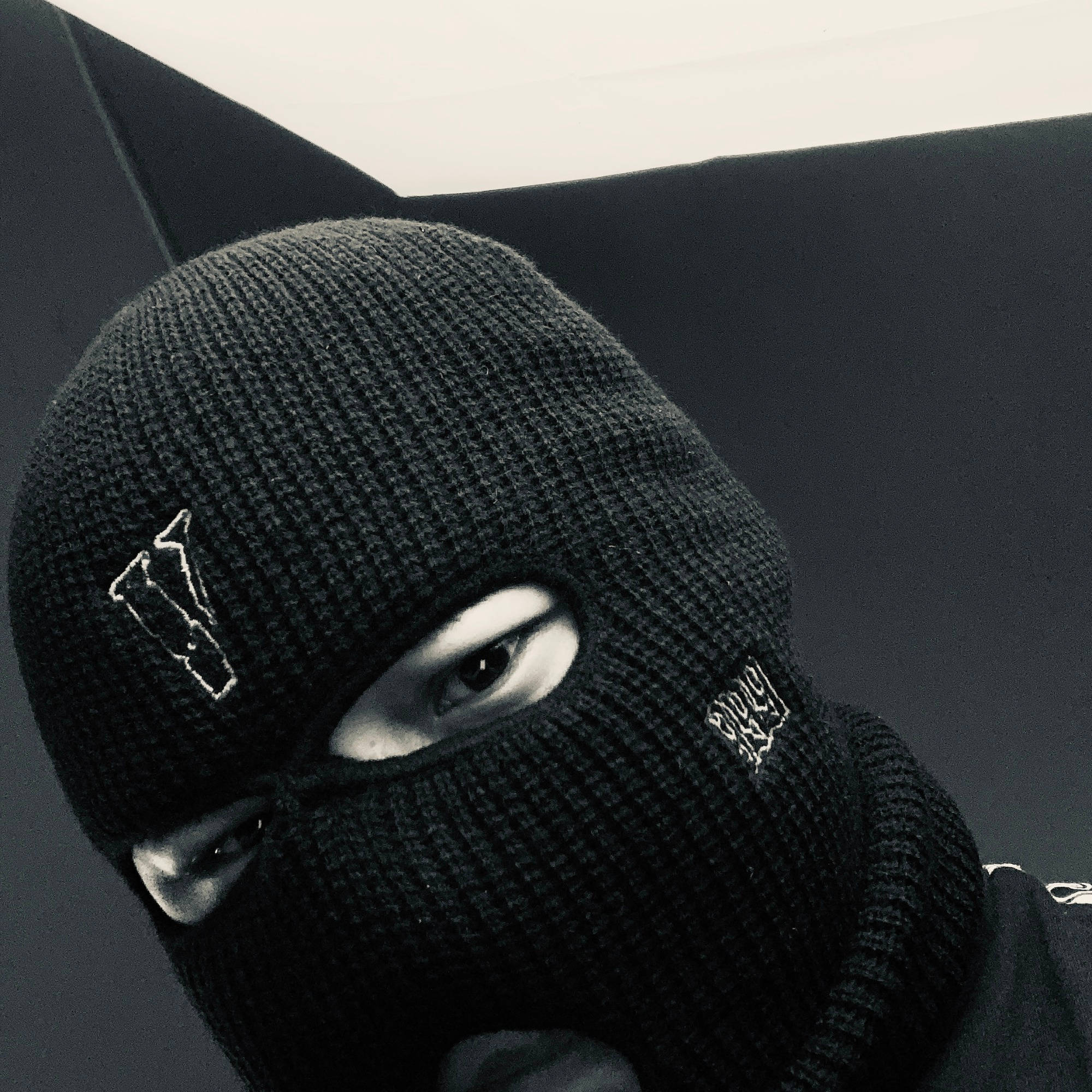 Vlone 桌布 壁紙 — 一個人戴著黑色針織巴拉克拉法帽，上面有白色刺繡標誌，從黑暗簡約的背景中強烈凝視。