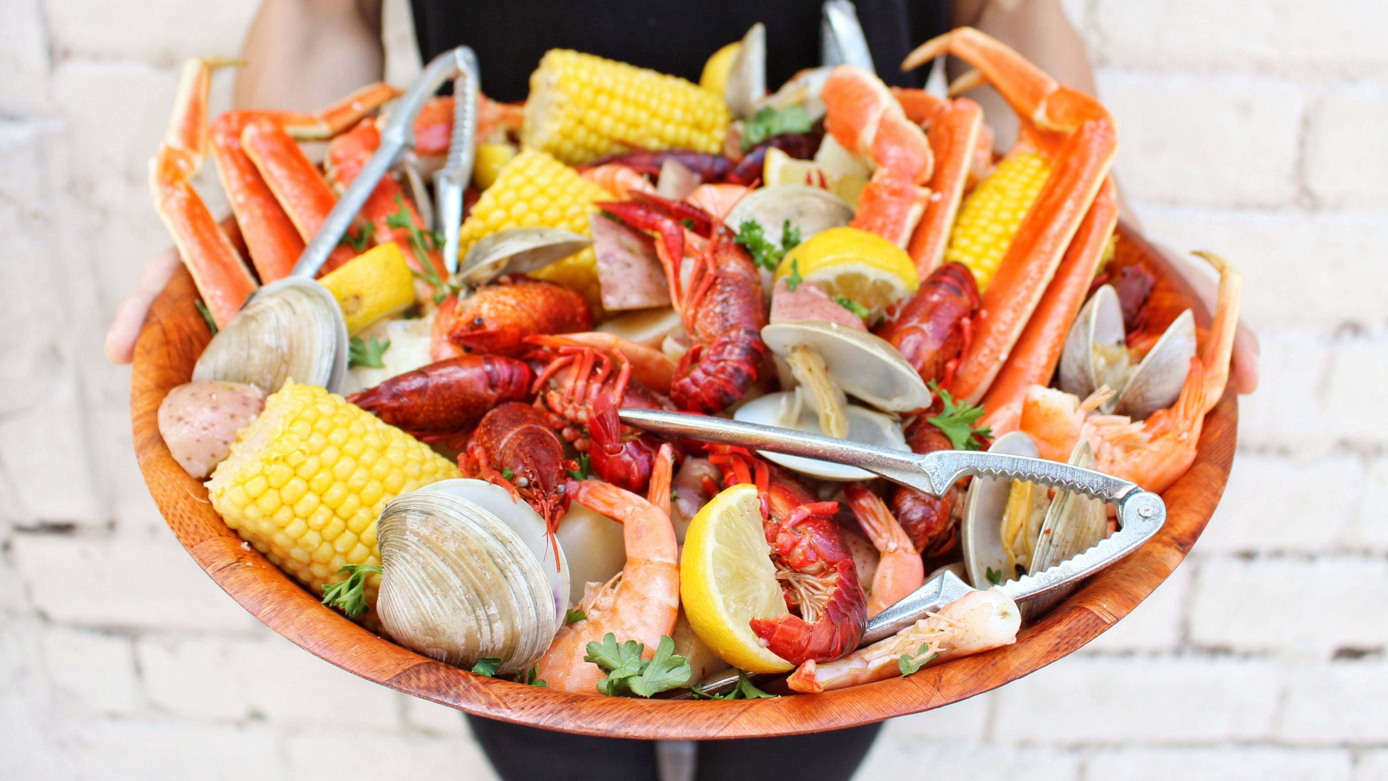 Seafood 桌布 壁紙 — 一個人手持一個木碗，裡面裝滿了豐富的海鮮煮物，包括小龍蝦、玉米、蛤蜊和檸檬角。