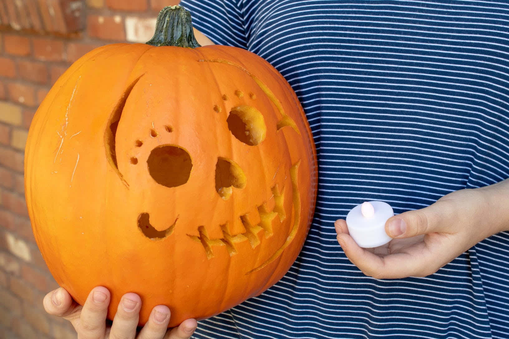 Pumpkin 桌布 壁紙 — 一個人手持一個雕刻過的橙色南瓜，帶有縫合的嘴巴和眼窩，同時點燃一支白色小蠟燭。