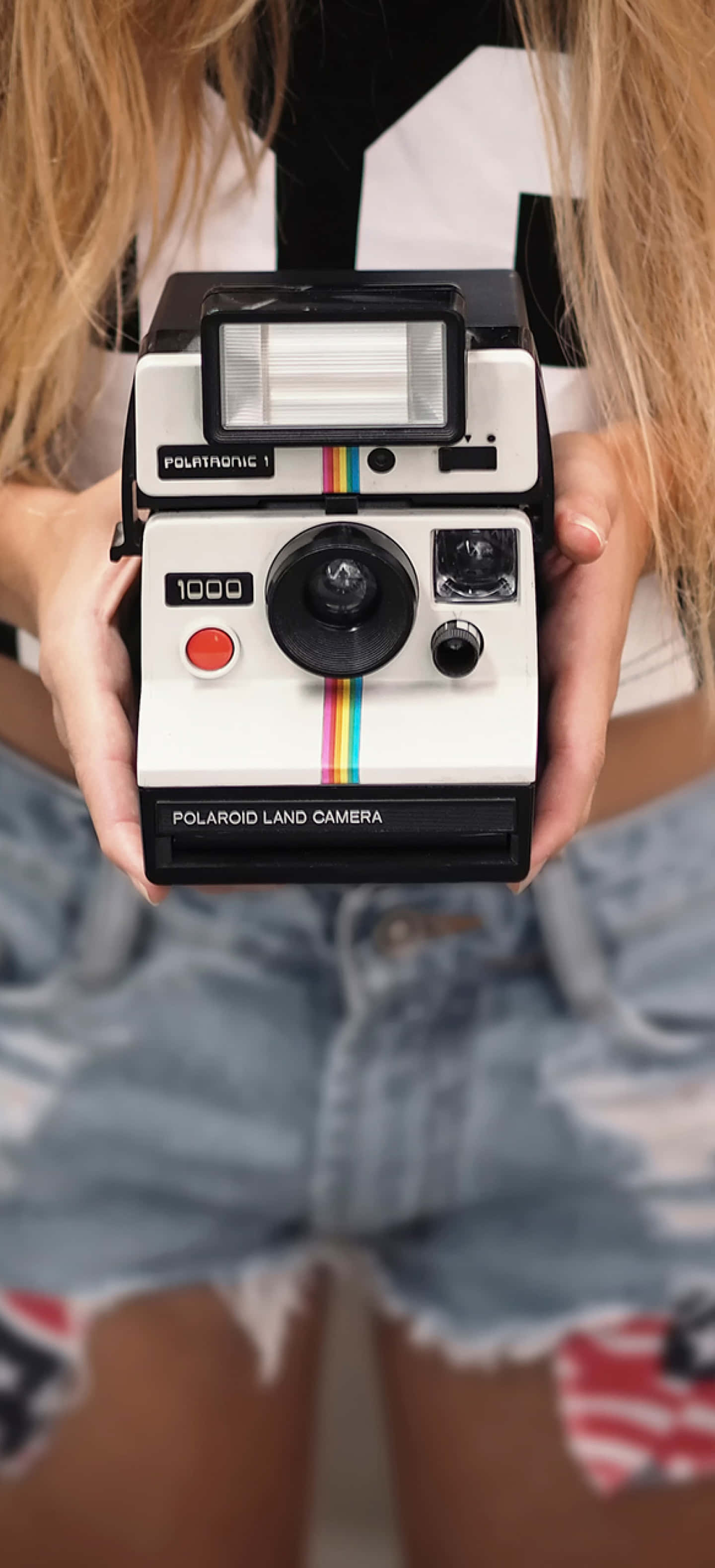 Photograph 桌布 壁紙 — 一個人手持一台復古Polaroid Land Camera 1000，背景是模糊的牛仔短褲和金髮。