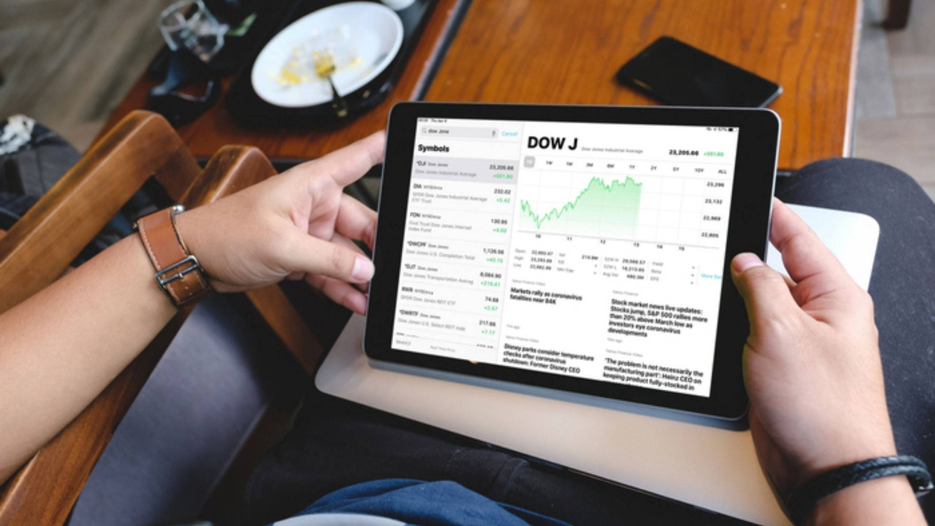 Dow Jones 桌布 壁紙 — 一個人手持平板電腦，顯示綠色上升的股票圖表及道瓊斯數據，坐在木桌旁，桌上有一盤食物。