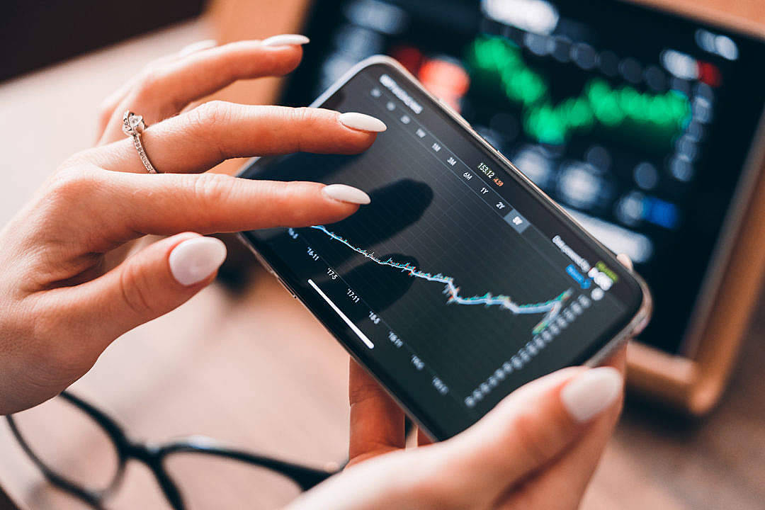 money iPhone 桌布 壁紙 — 一個人手持智慧型手機，顯示帶有綠色和藍色線條的股票市場圖表，背景模糊地呈現一個更大的螢幕和眼鏡。