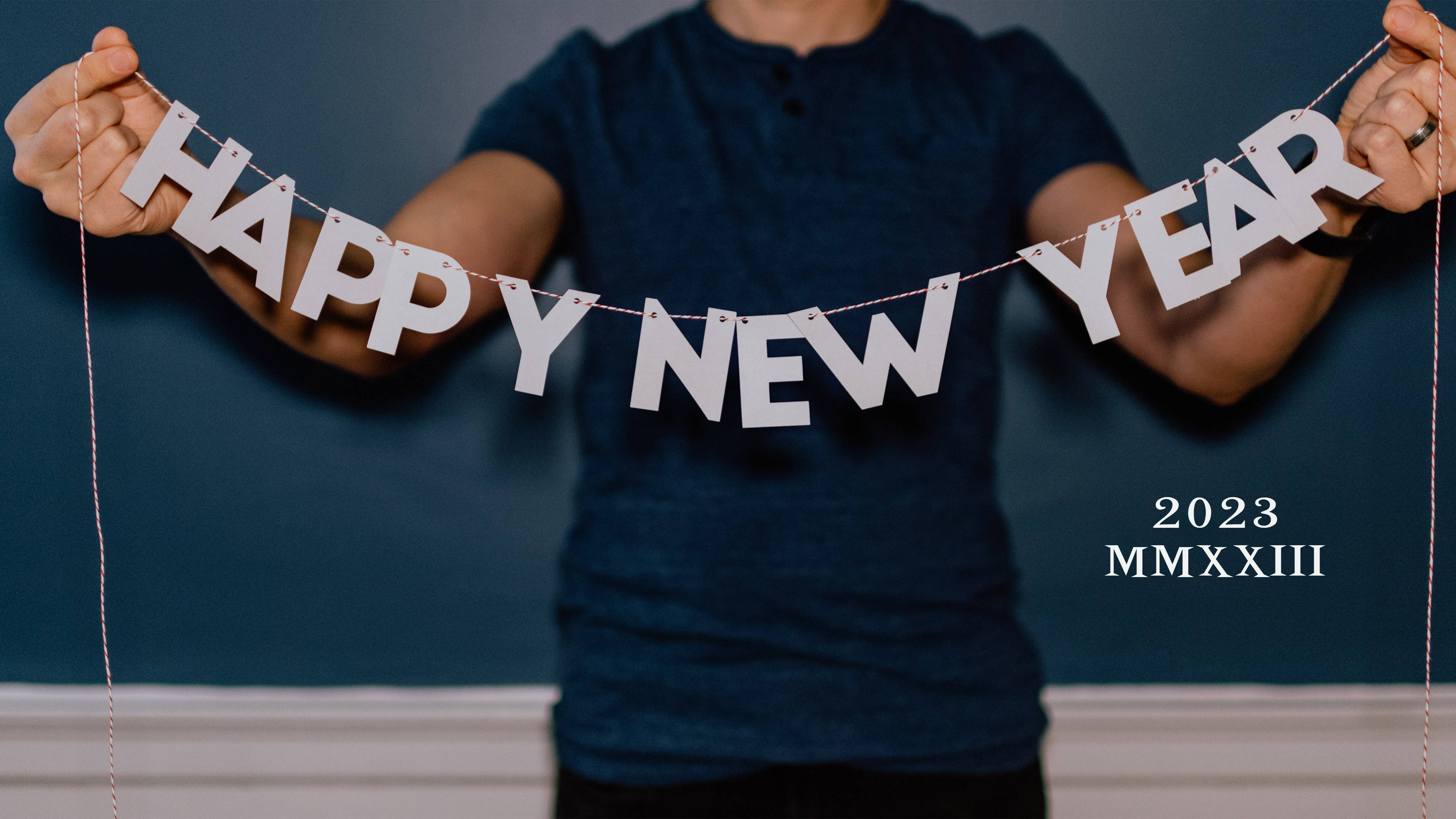 New Year 桌布 壁紙 — 一個人手持白色「HAPPY NEW YEAR」橫幅，背景為深藍色牆壁，下方有「2023 MMXXIII」文字。