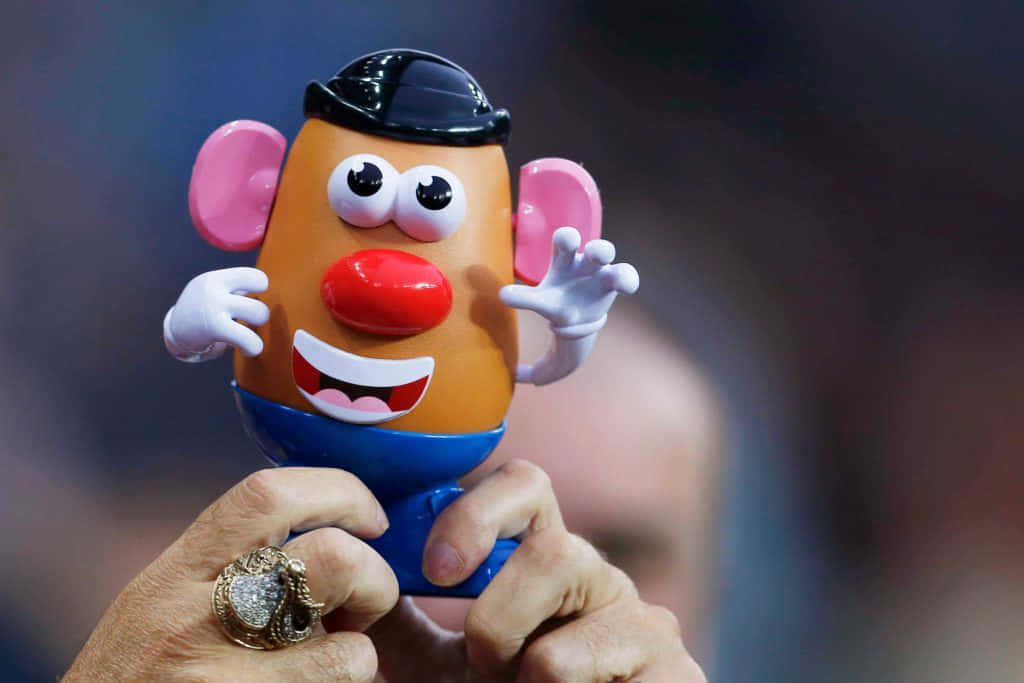 Mr Potato Head 桌布 壁紙 — 一個人手持經典Mr Potato Head玩具的特寫，玩具擁有粉紅色耳朵、紅色鼻子和藍色褲子，背景模糊。
