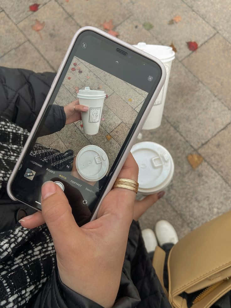 iPhone 美學 桌布 壁紙 — 一個人手持電話拍攝散落秋葉的人行道上的一杯咖啡。