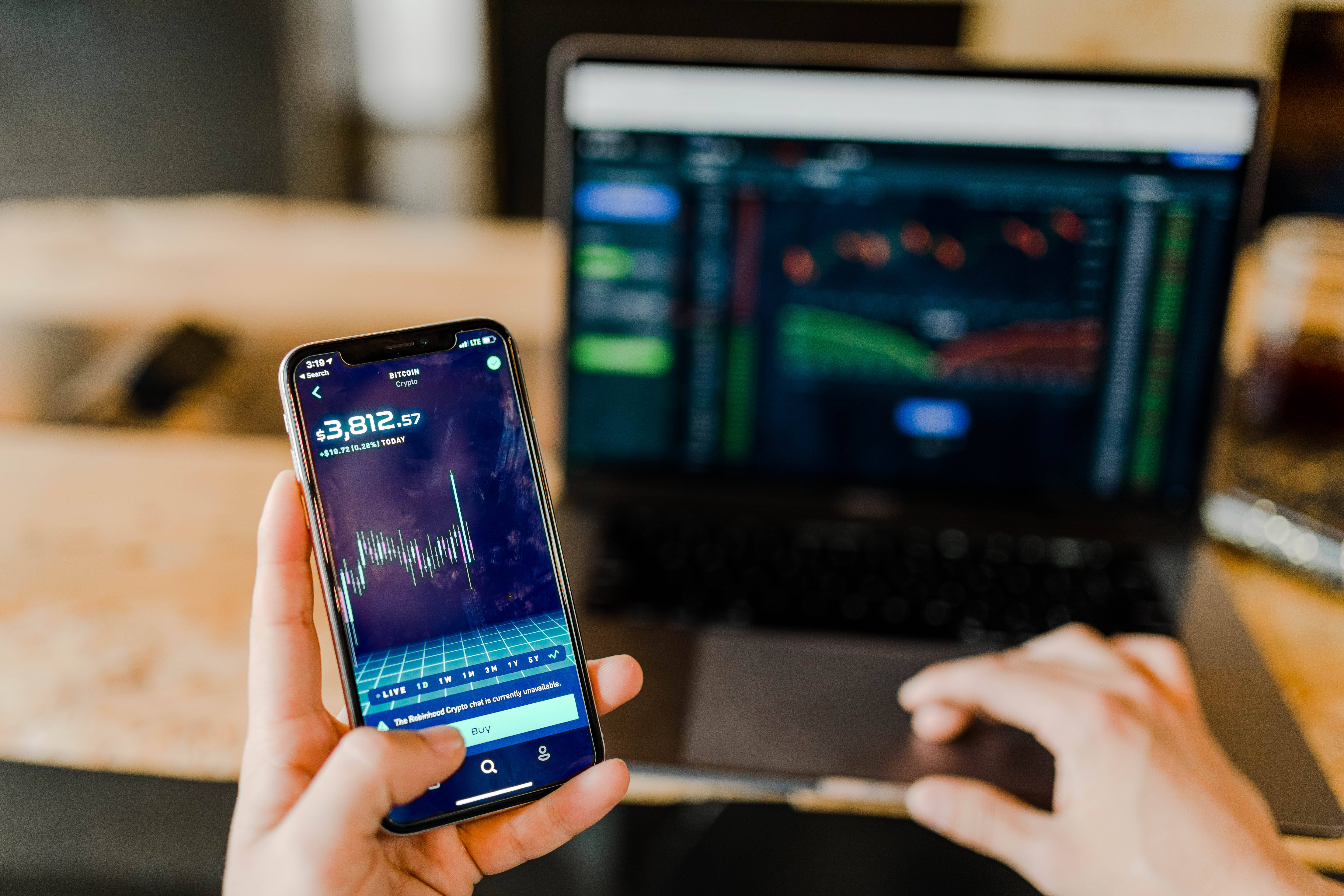 Cryptocurrency 桌布 壁紙 — 一個人手持顯示加密貨幣交易應用程式的手機，同時在背景有金融圖表的筆記型電腦上打字。