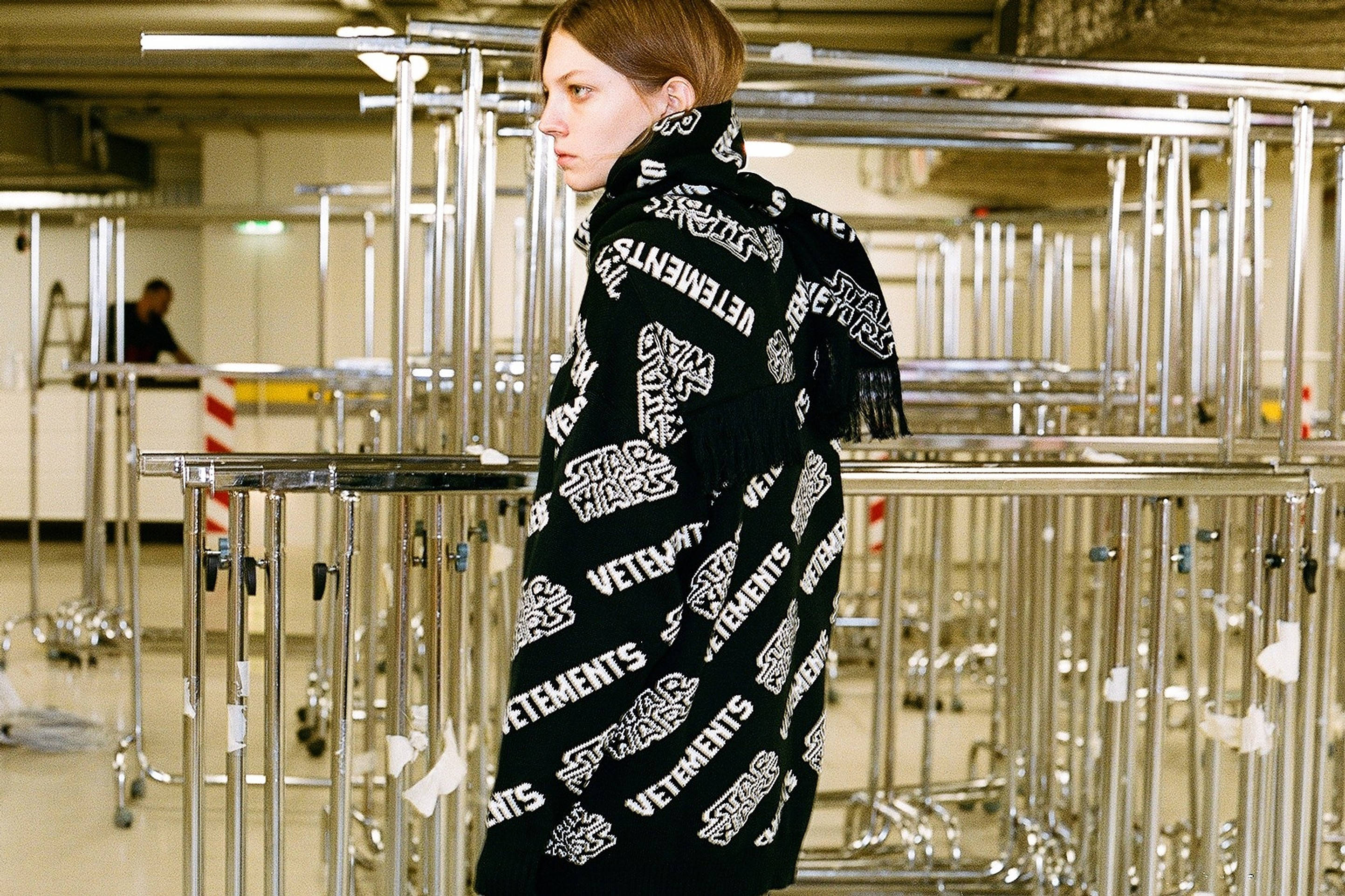 Vetements 桌布 壁紙 — 一個人披著黑色Vetements圍巾，上面有白色重複標誌，站在倉庫式空間的鉻製欄杆之間。