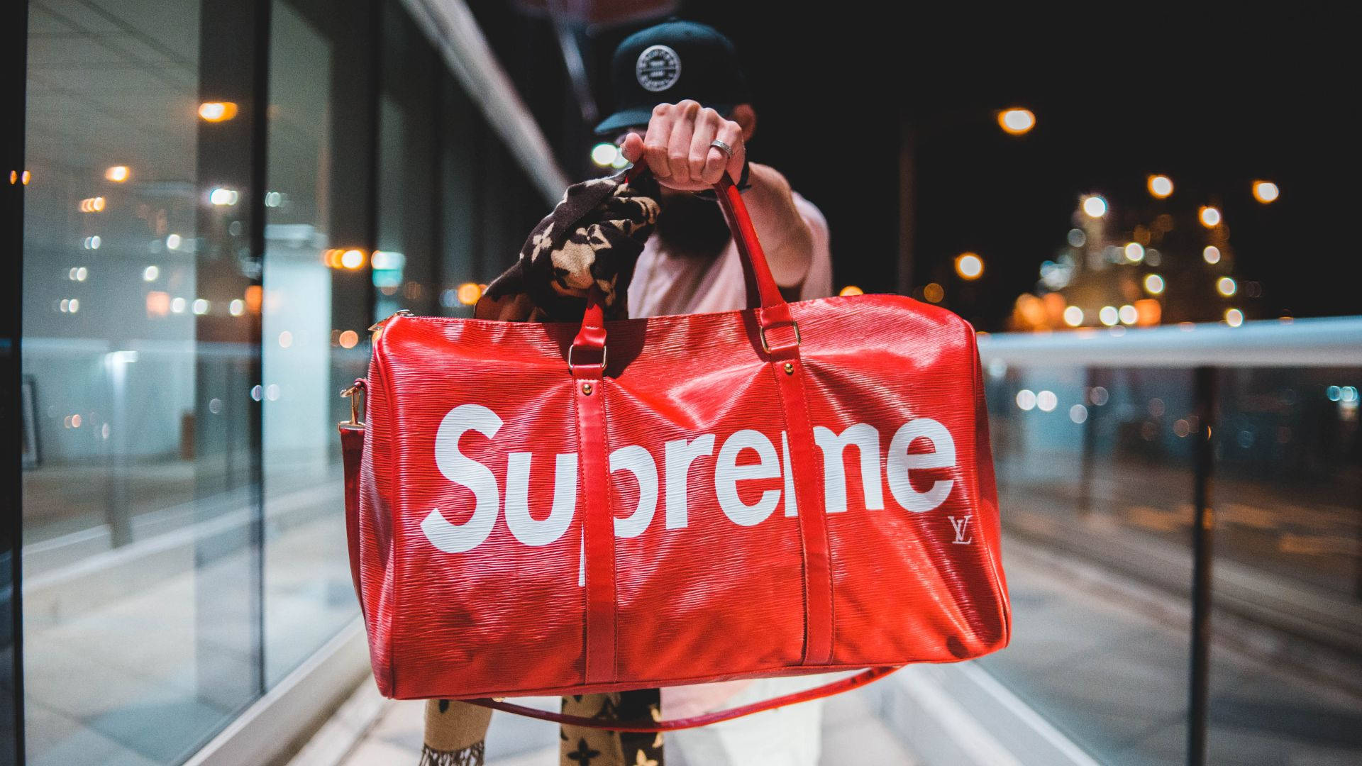 Cool Supreme 桌布 壁紙 — 一個人拿著一個帶有白色標誌的紅色Supreme行李袋，背景是模糊的城市夜景。