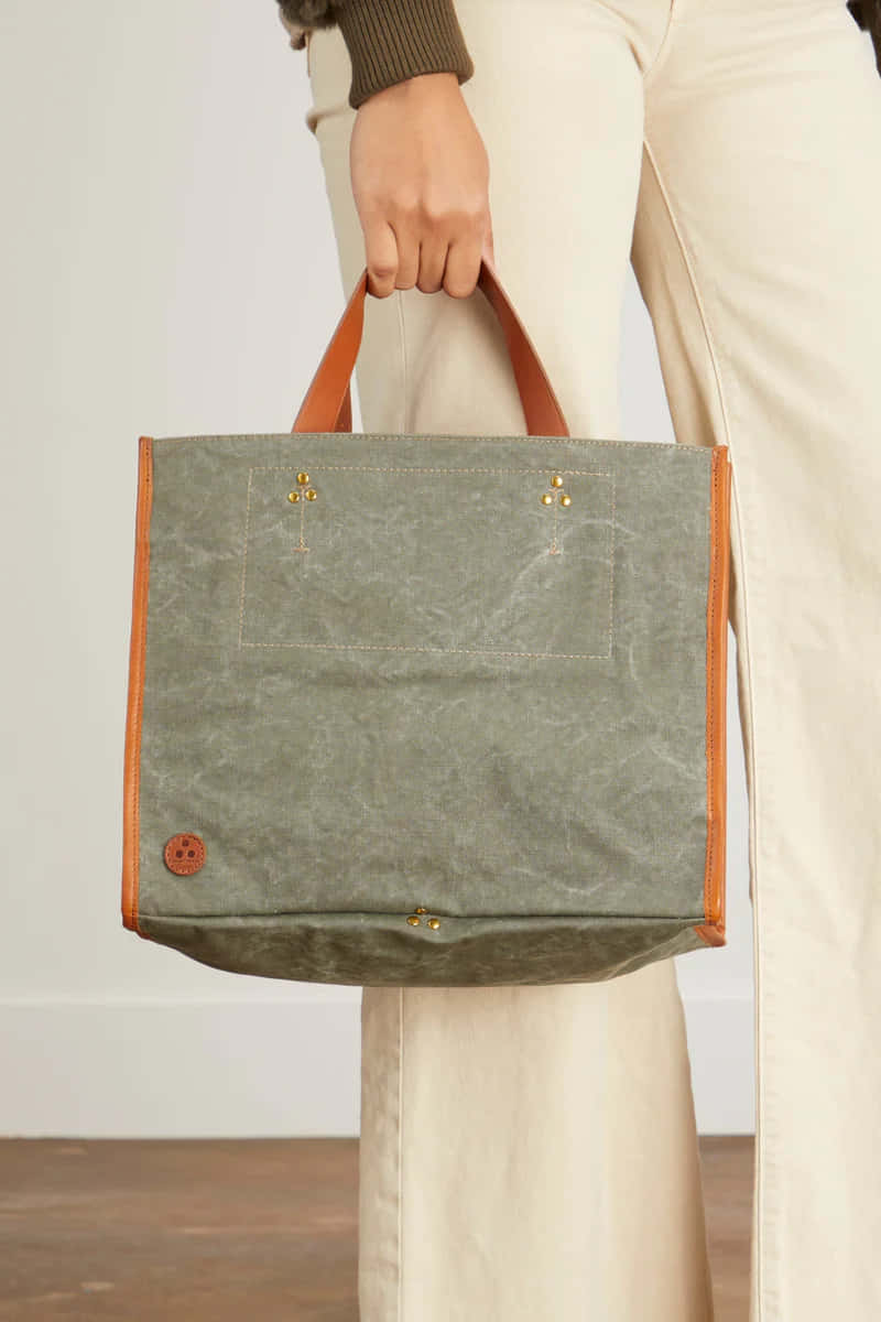 Handbag 桌布 壁紙 — 一個人拿著一個綠色蠟帆布手提袋，配有棕褐色皮革手柄和黃銅五金件，背景為簡約室內風格。