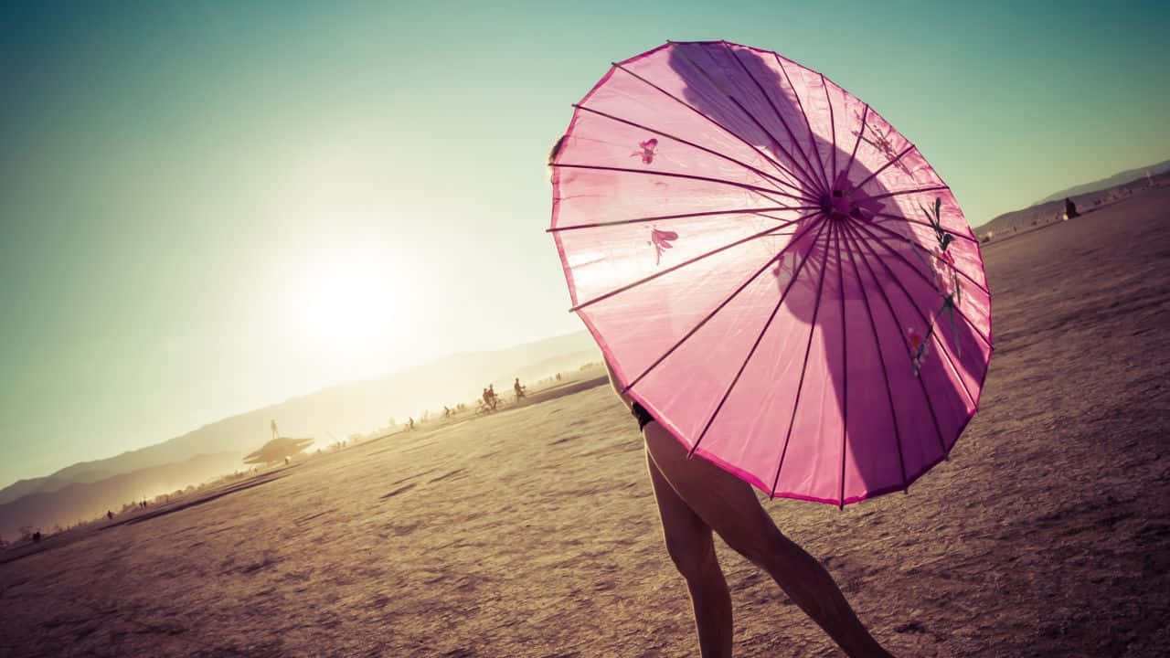 Beach Umbrella 桌布 壁紙 — 一個人拿著一把粉紅色遮陽傘，遮住他們的臉，對著明亮的沙漠陽光和遠山。
