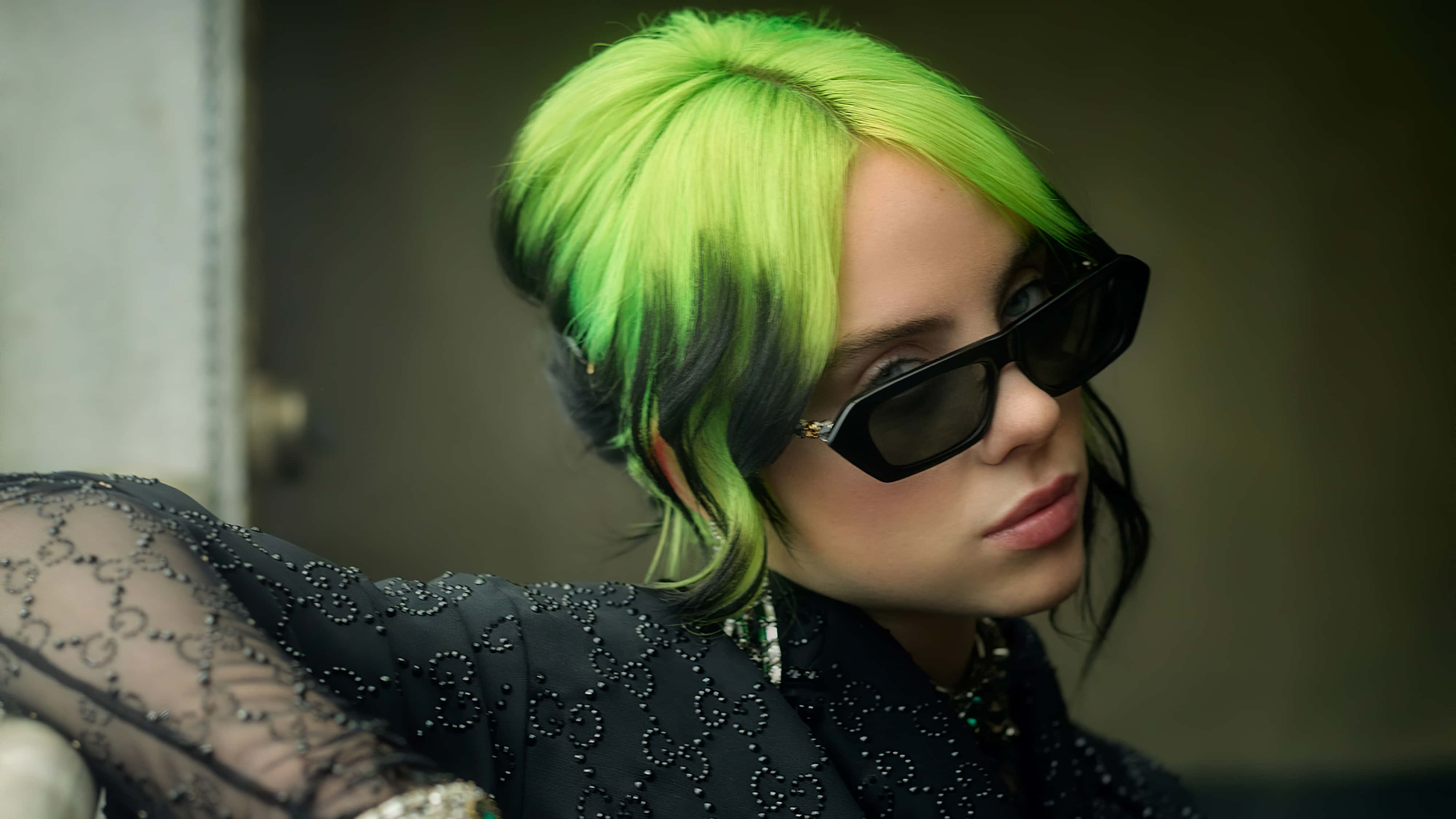 Billie Eilish Laptop 桌布 壁紙 — 一個人擁有充滿活力的綠色頭髮和黑色太陽眼鏡，穿著黑色珠飾服裝，背景柔和。