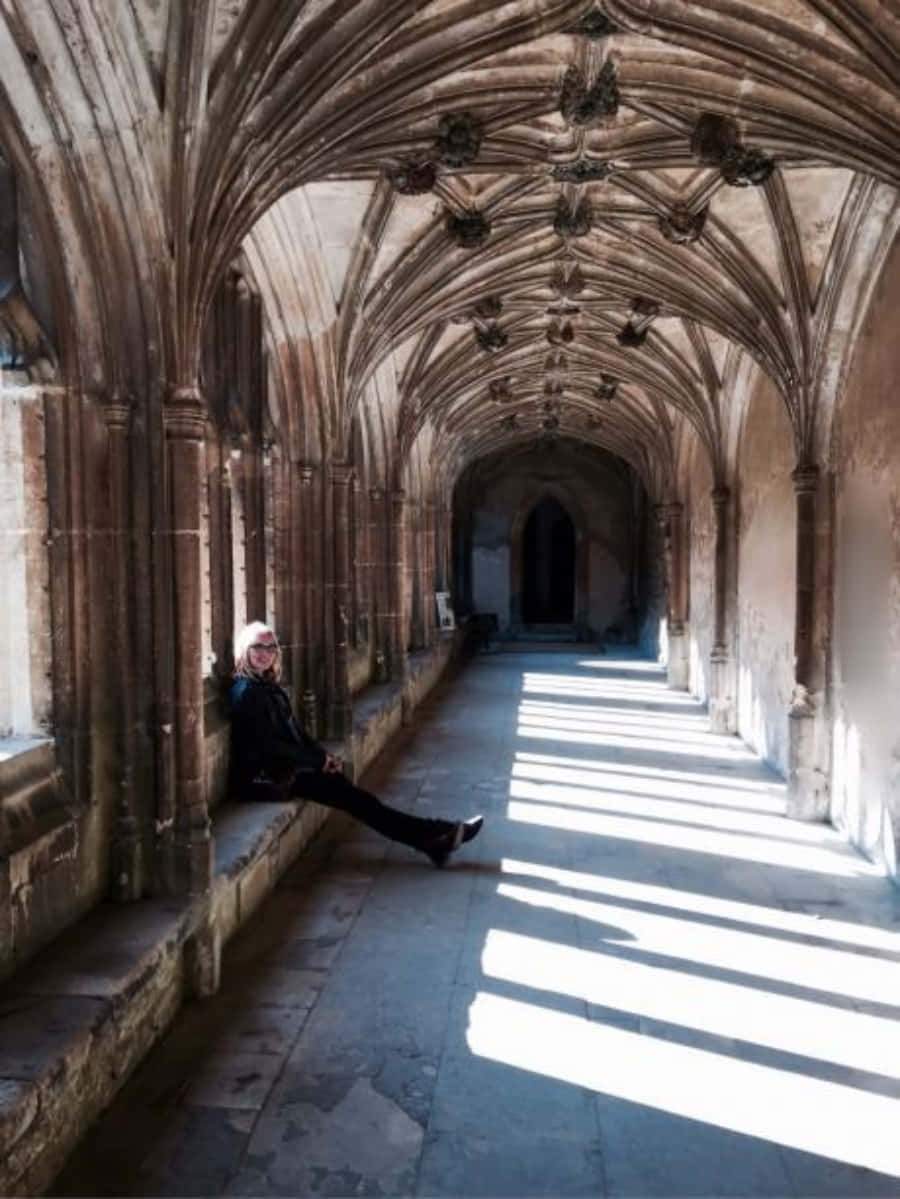 The Hogwarts Forbidden Corridor 桌布 壁紙 — 一個人放鬆地坐在石凳上，身處陽光普照的哥德式迴廊，擁有精緻的拱形天花板和拱形窗戶。