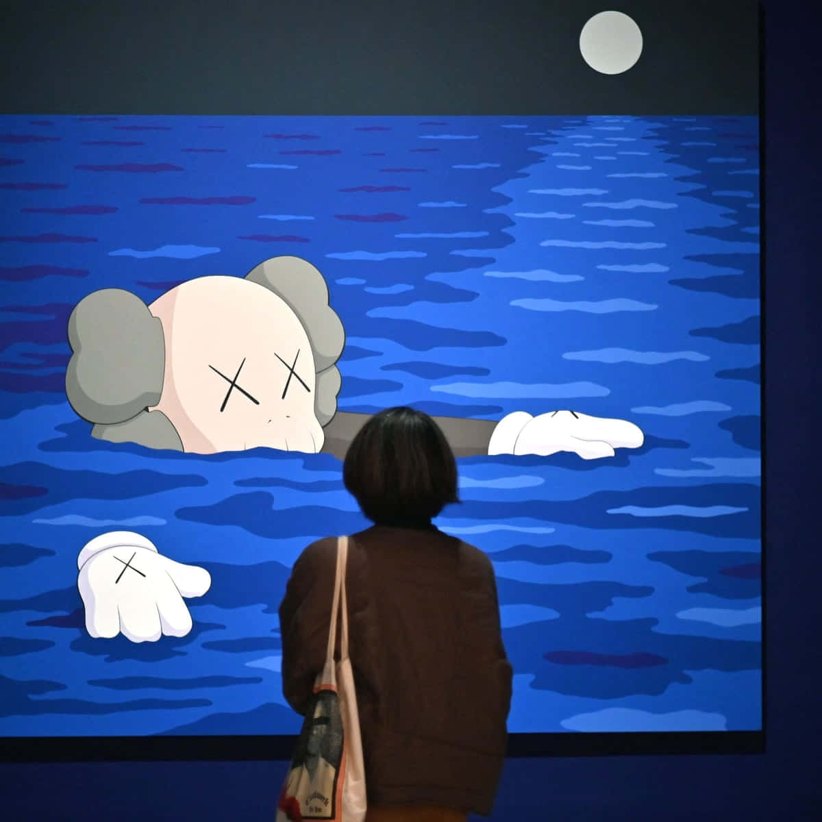 KAWS 桌布 壁紙 — 一個人正在觀看一個大螢幕，顯示一個卡通風格的KAWS角色在滿月下浸在藍色水中。